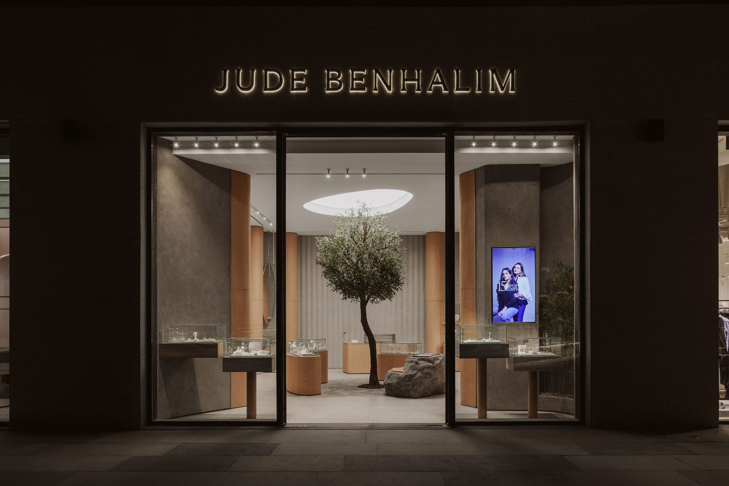 Jude Benhalim luxury jewelry boutique storefront at night with warm lighting and olive tree centerpiece display.