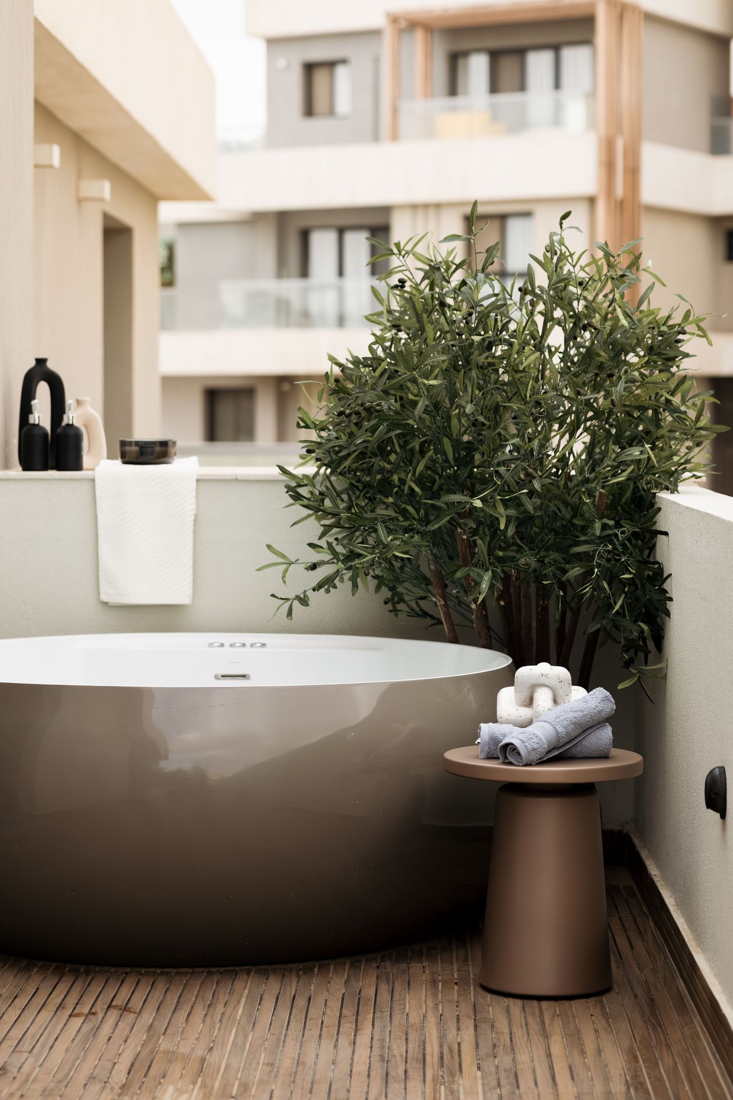 Round freestanding outdoor bathtub on wooden deck with green plant and folded towels.
