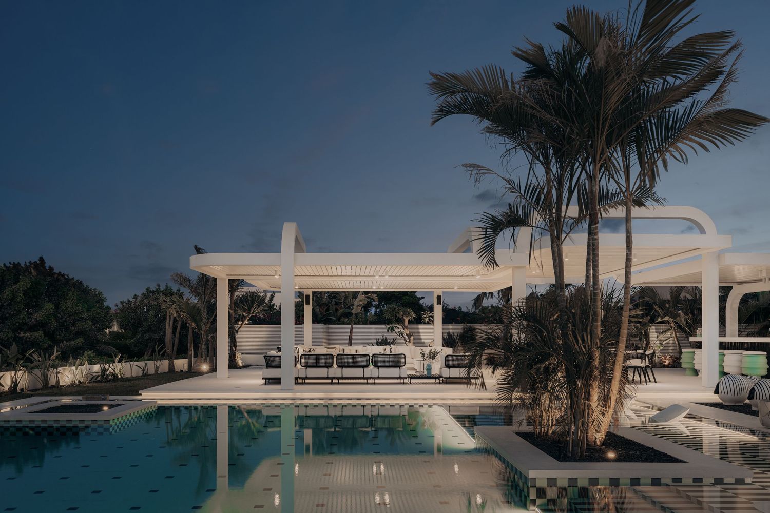 Luxury outdoor pool area with white pergola, palm trees, and elegant seating at dusk.