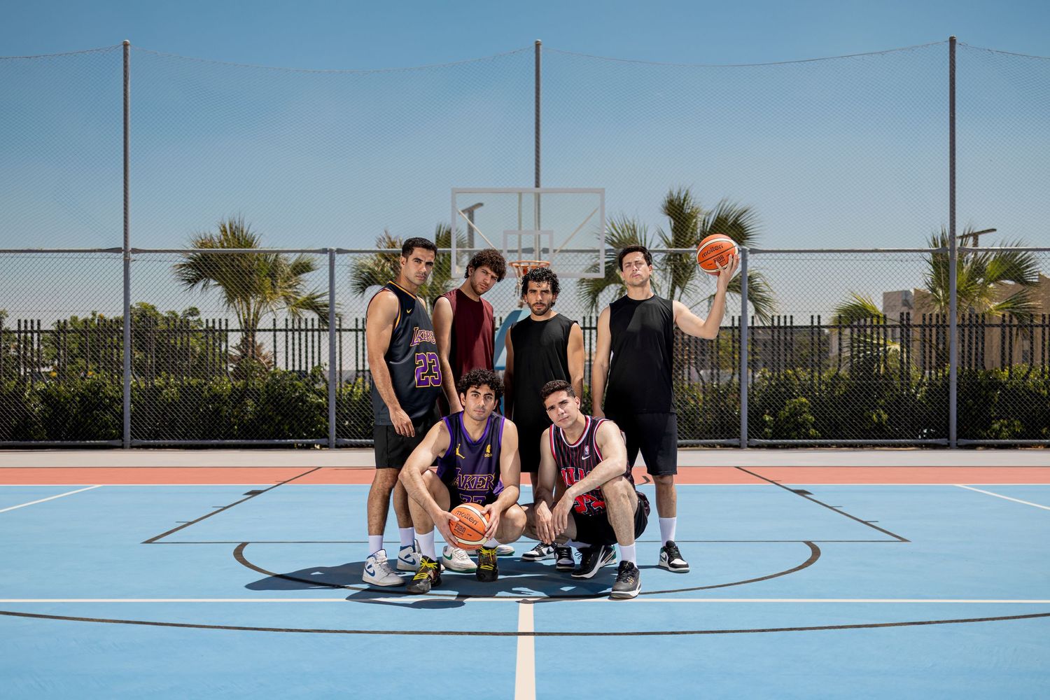 Group of five teenage basketball players posing together on an outdoor blue basketball court on a sunny day.