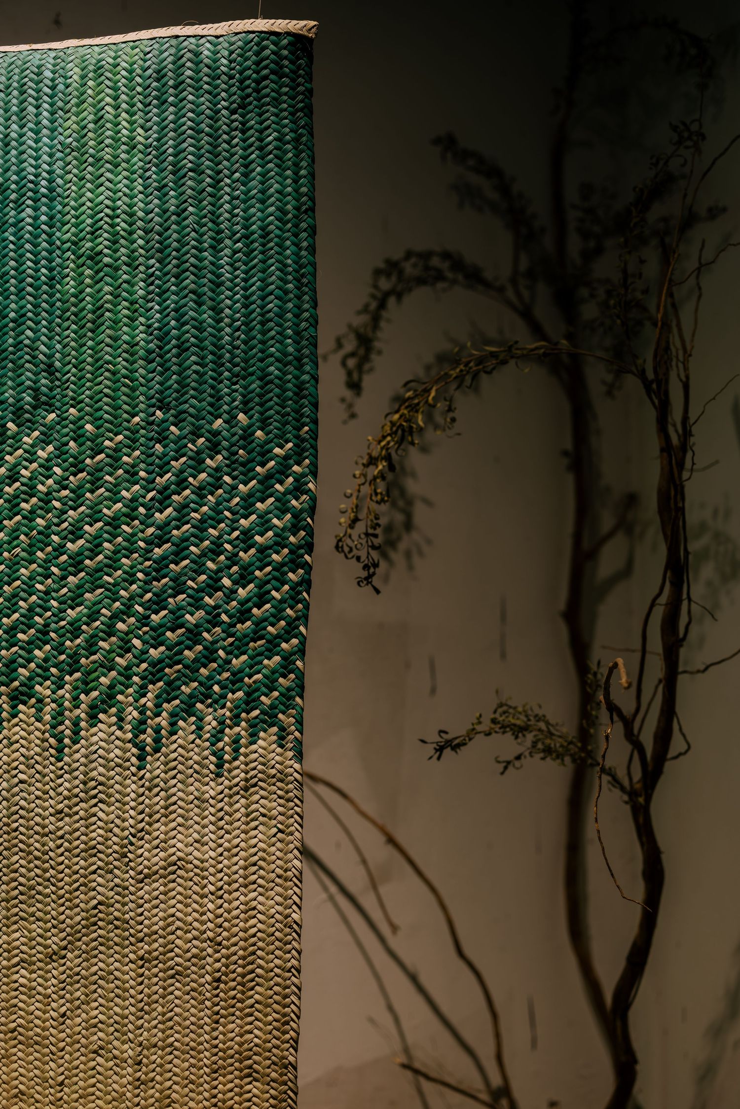 Green and gold woven textile wall hanging beside dried branches casting shadows on a dark wall.