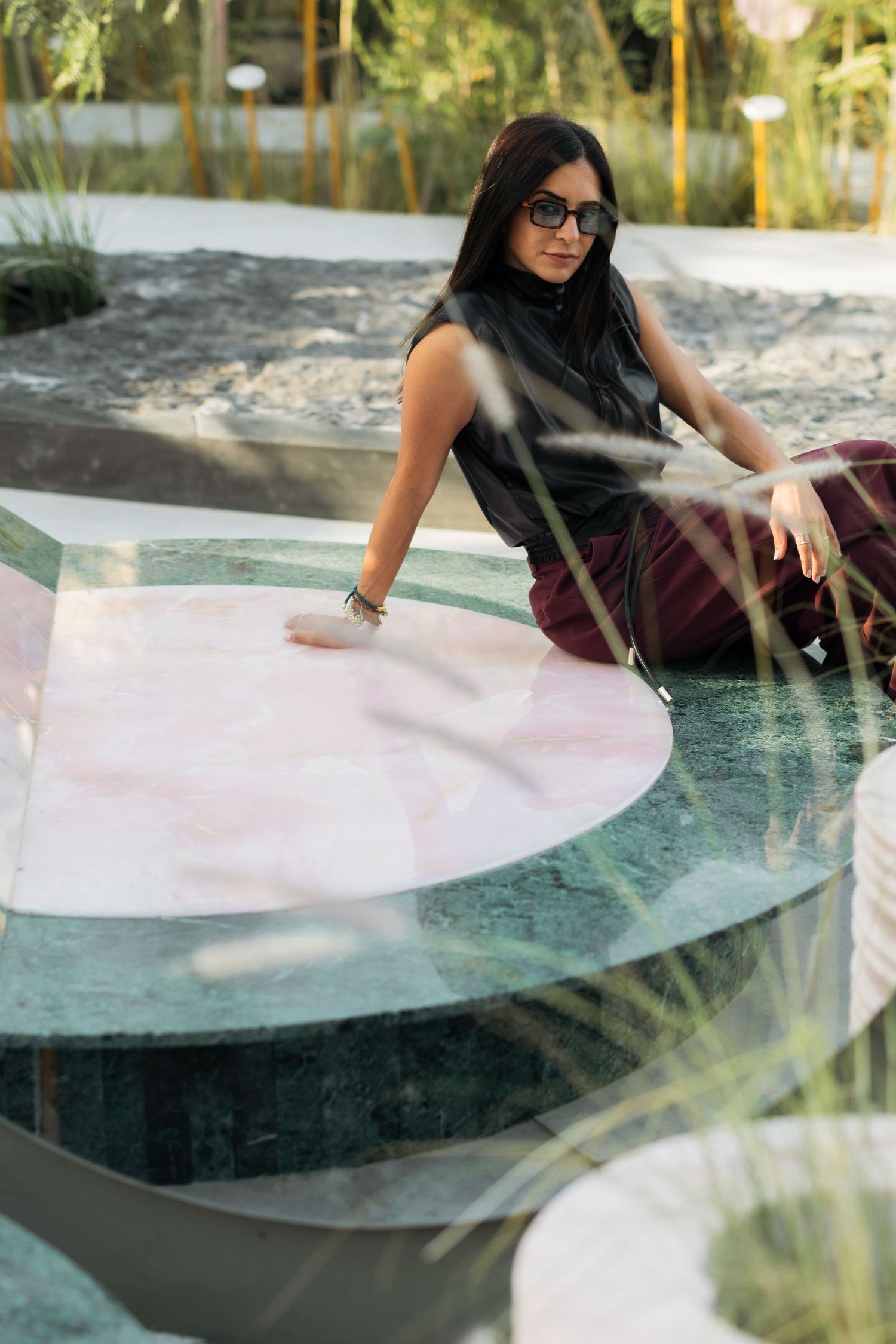Woman in sunglasses and dark outfit sitting on a large green and pink marble circular installation outdoors.