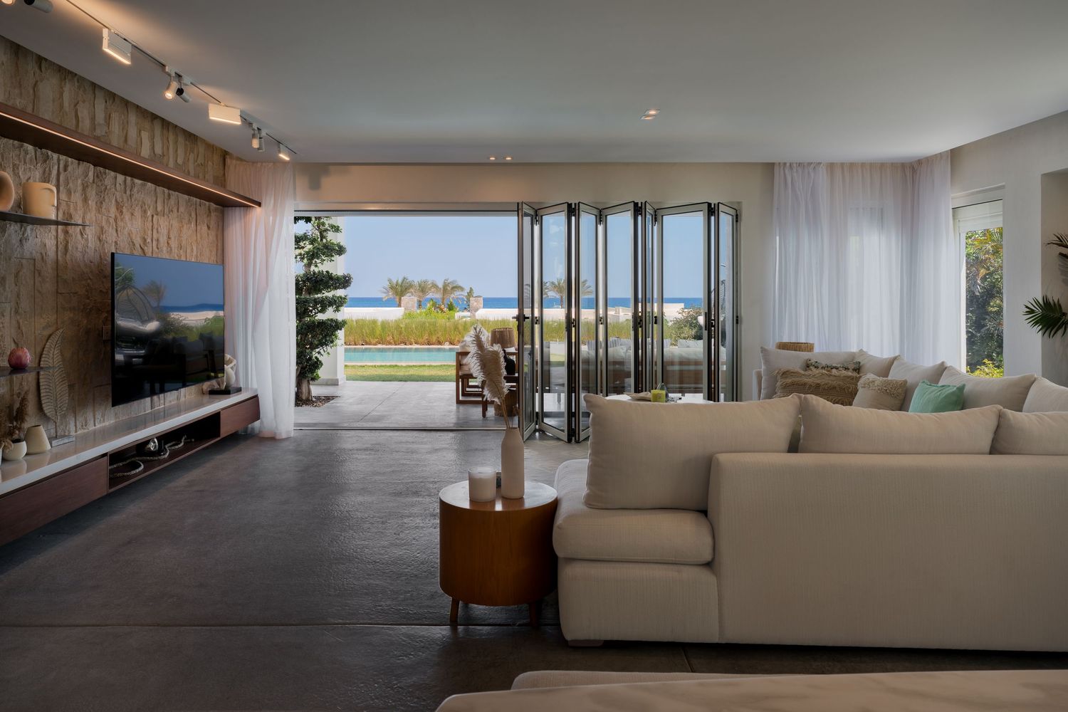 Bright modern living room with beige sofas, dark tile floors, and open bifold doors revealing a scenic ocean view.