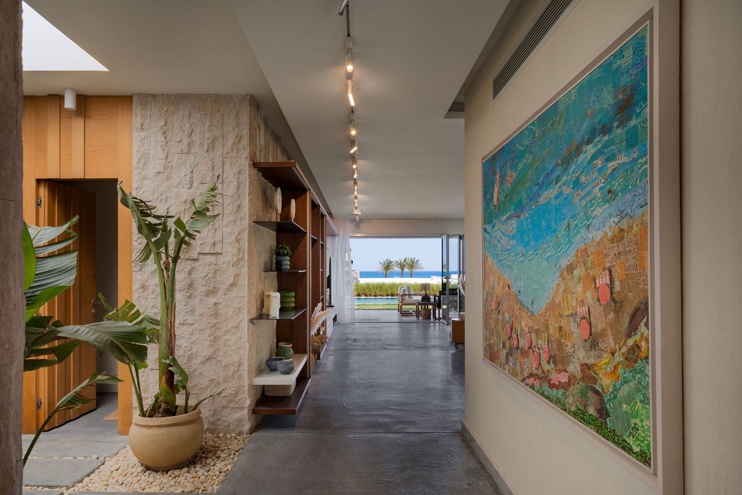 Modern villa corridor with polished concrete floors, track lighting, stone walls, tropical plants, and colorful abstract artwork.