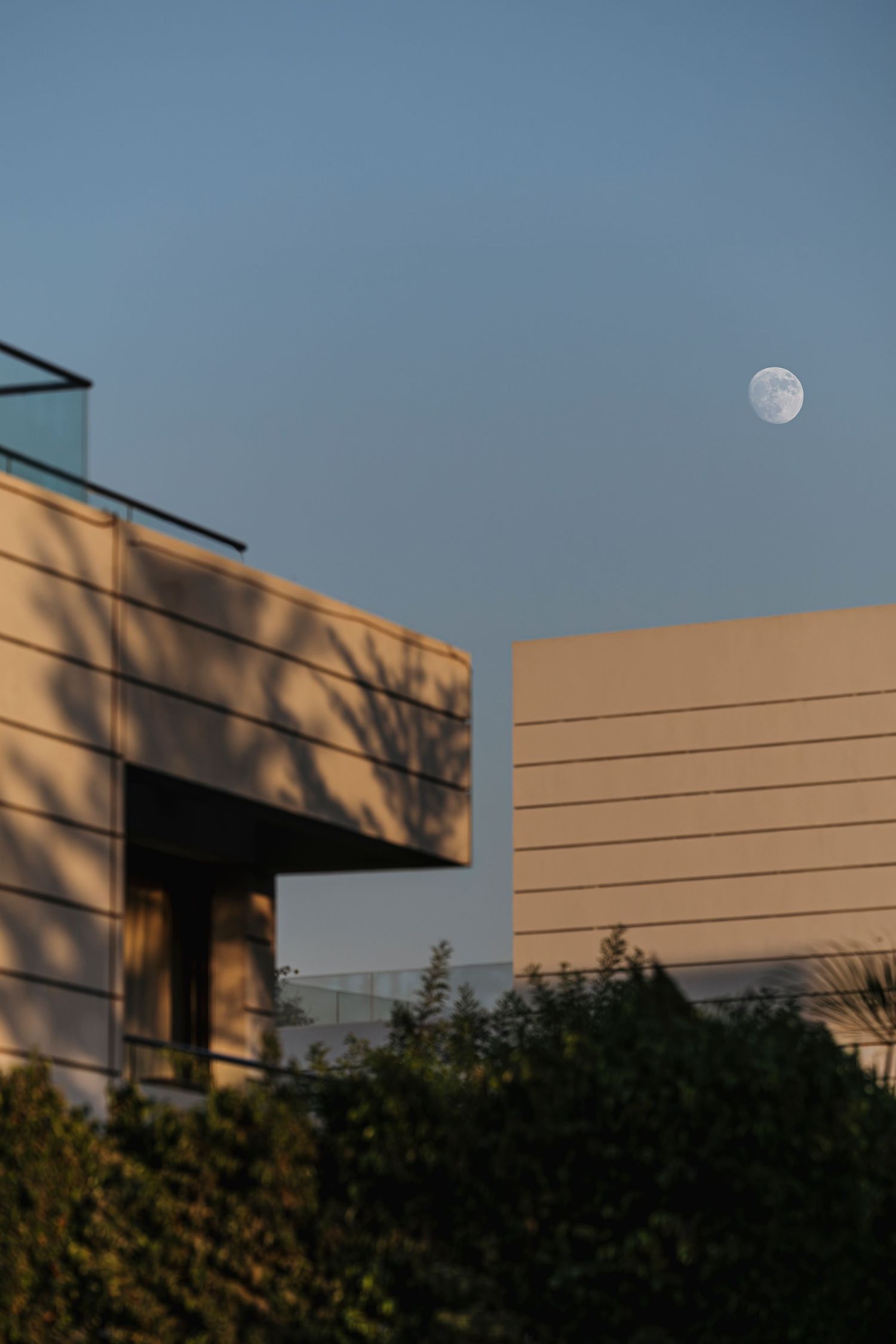 Modern building facades at golden hour with a half moon visible in the clear blue sky above lush green shrubs.