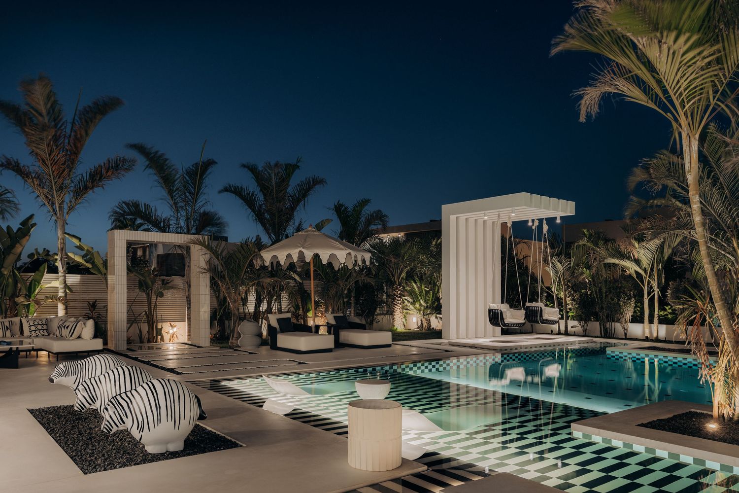 Luxury outdoor pool area at night with palm trees, white cabanas, and a checkered patio under a dark blue sky.