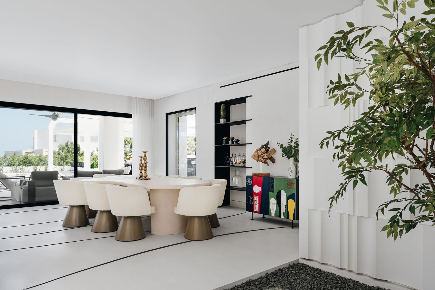 Modern white dining room with cream chairs, oval table, built-in shelving, and indoor tree beside floor-to-ceiling windows.