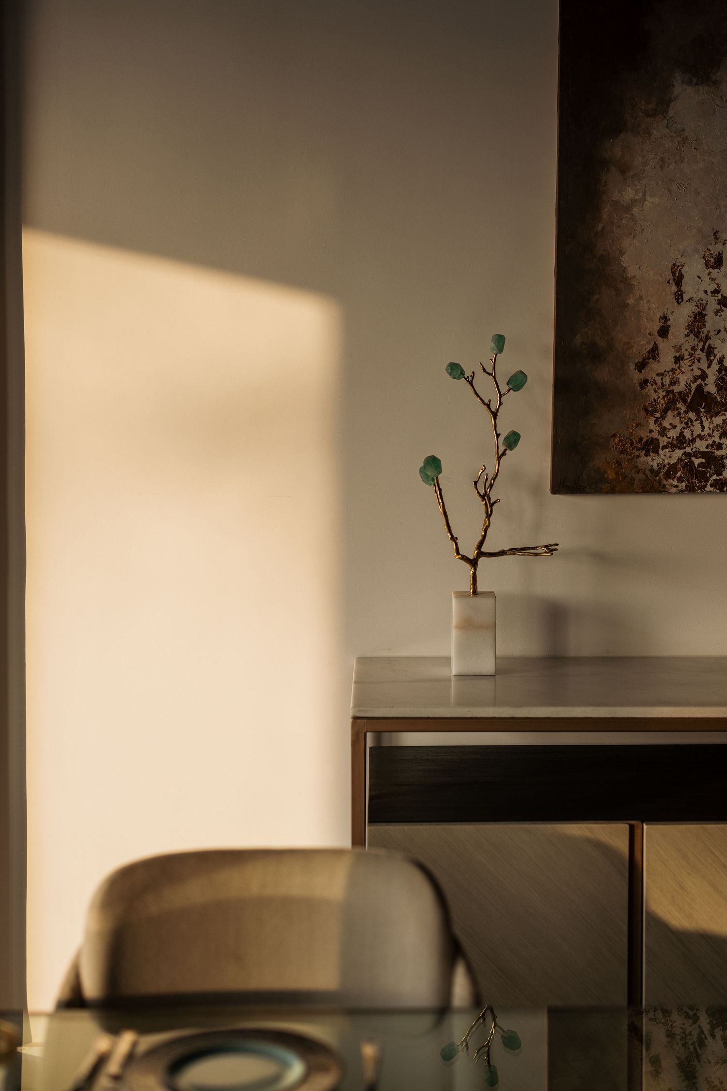 Minimalist console table with a delicate branch vase casting warm shadows on a neutral wall beside abstract dark artwork.