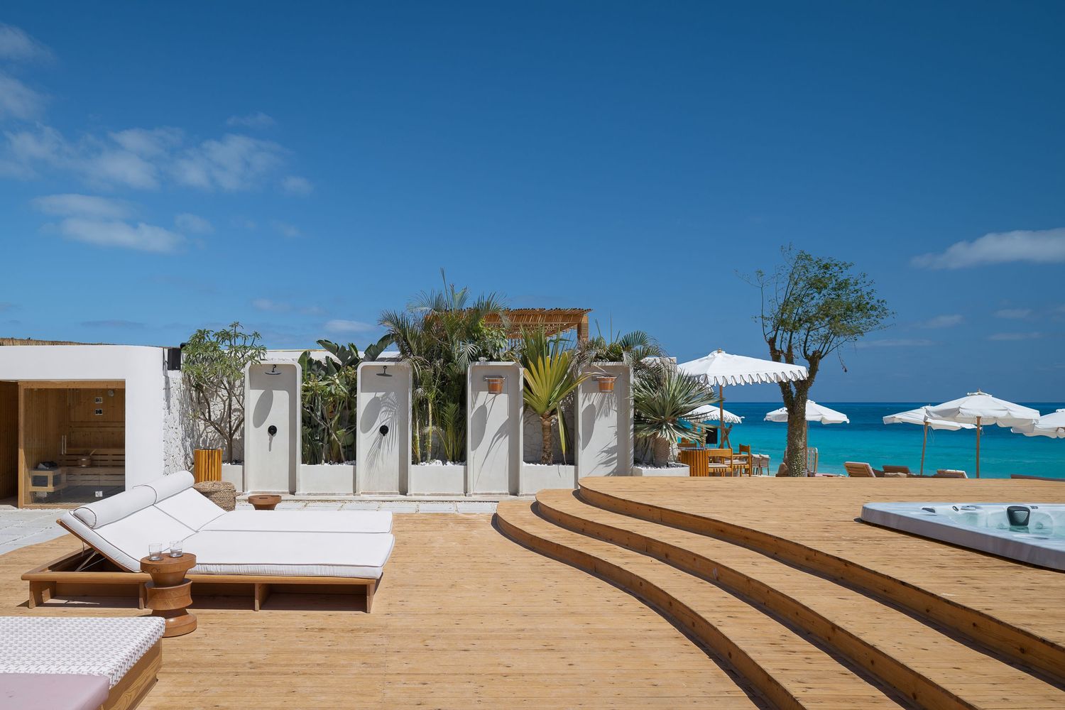 Luxury beachfront deck with white sun loungers, outdoor showers, tropical plants, and turquoise ocean views under blue sky.