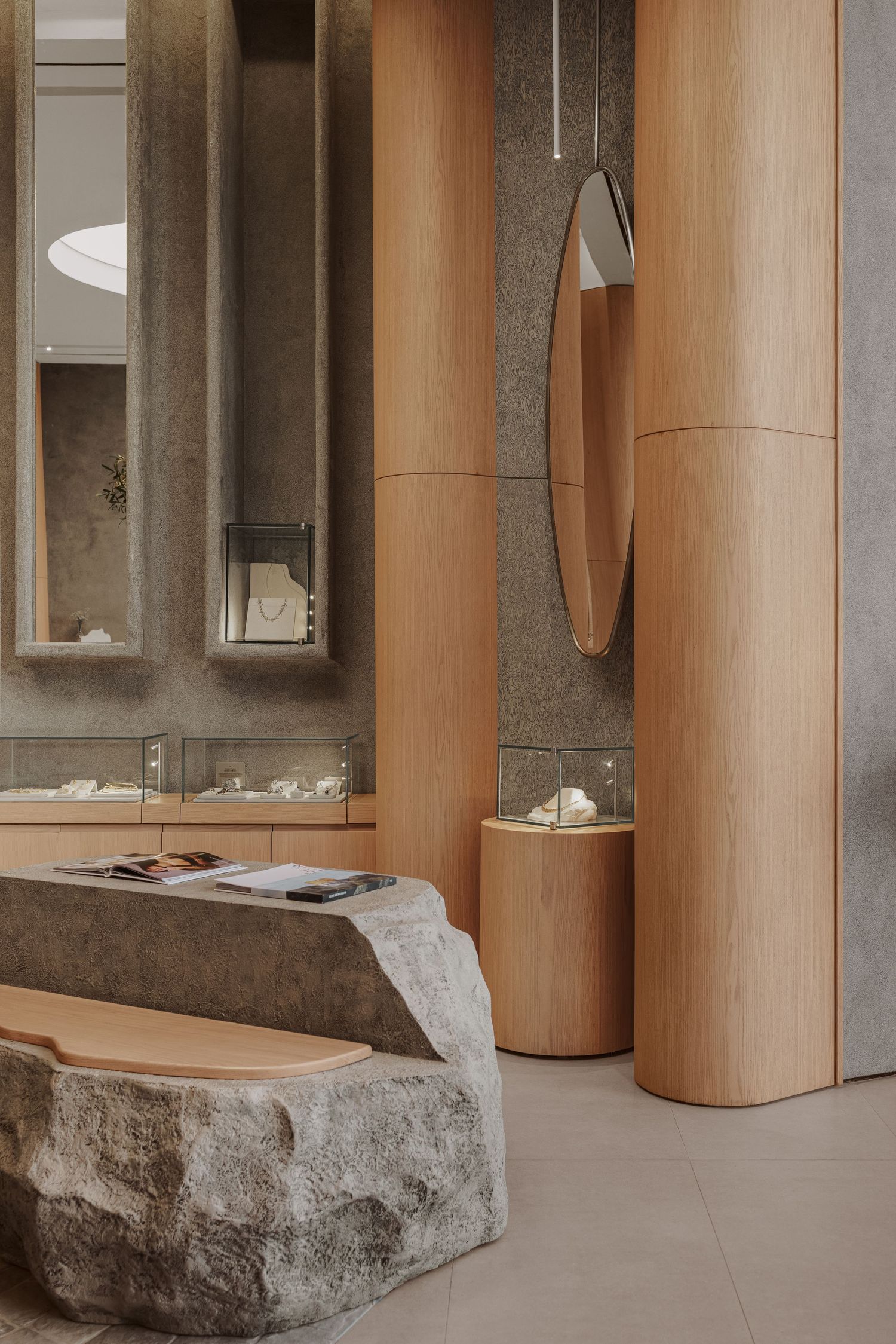 Upscale jewelry store interior featuring cylindrical wood columns, oval mirror, stone table, and glass display cases.