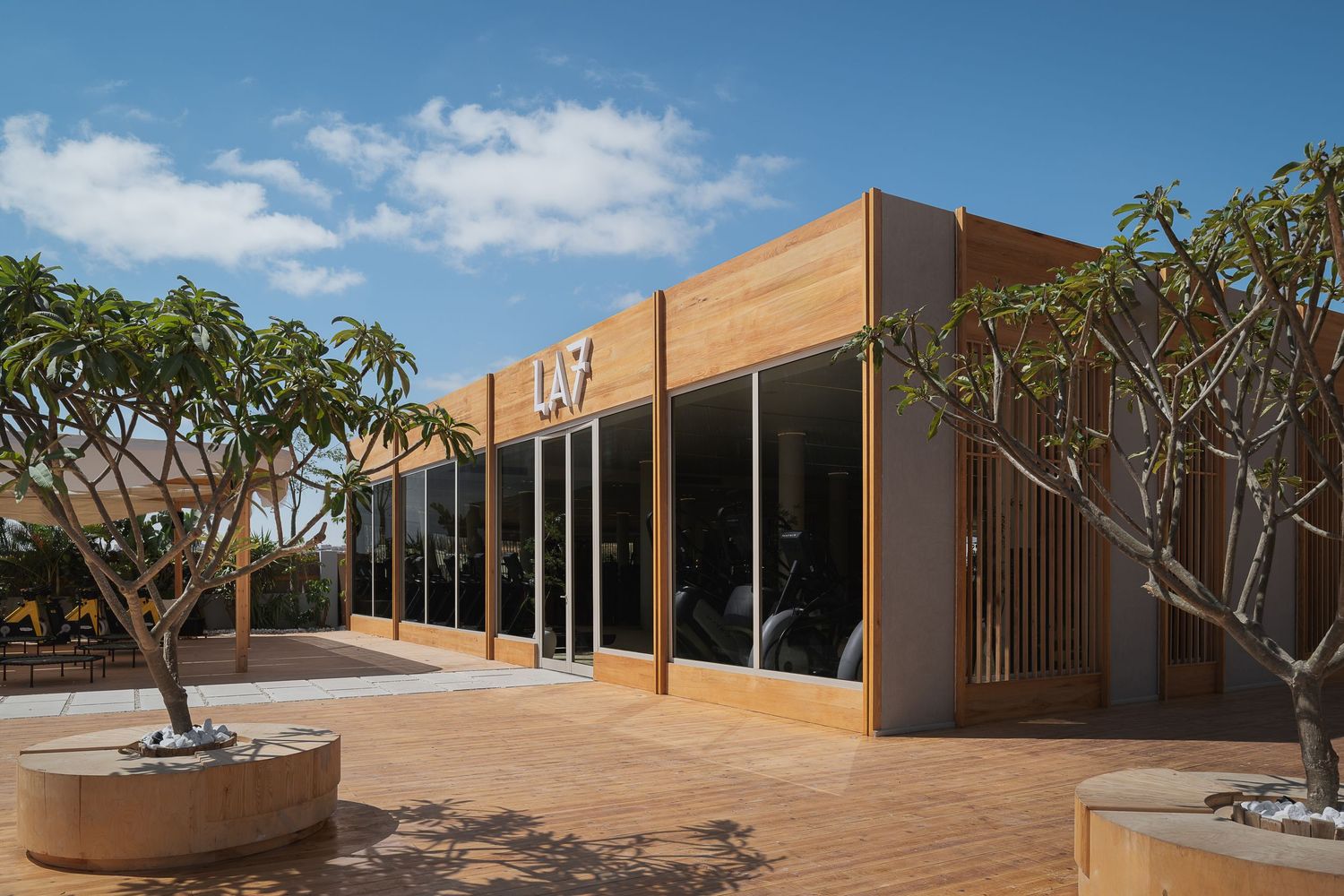 LA7 fitness center exterior with timber-clad facade, large glass windows, and native trees in sunny courtyard.