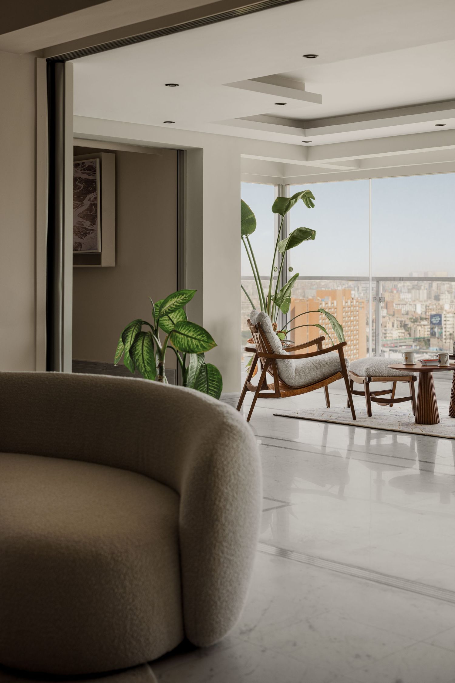 Contemporary living space with curved boucle sofa, marble floors, tropical plants, and city views through large windows.