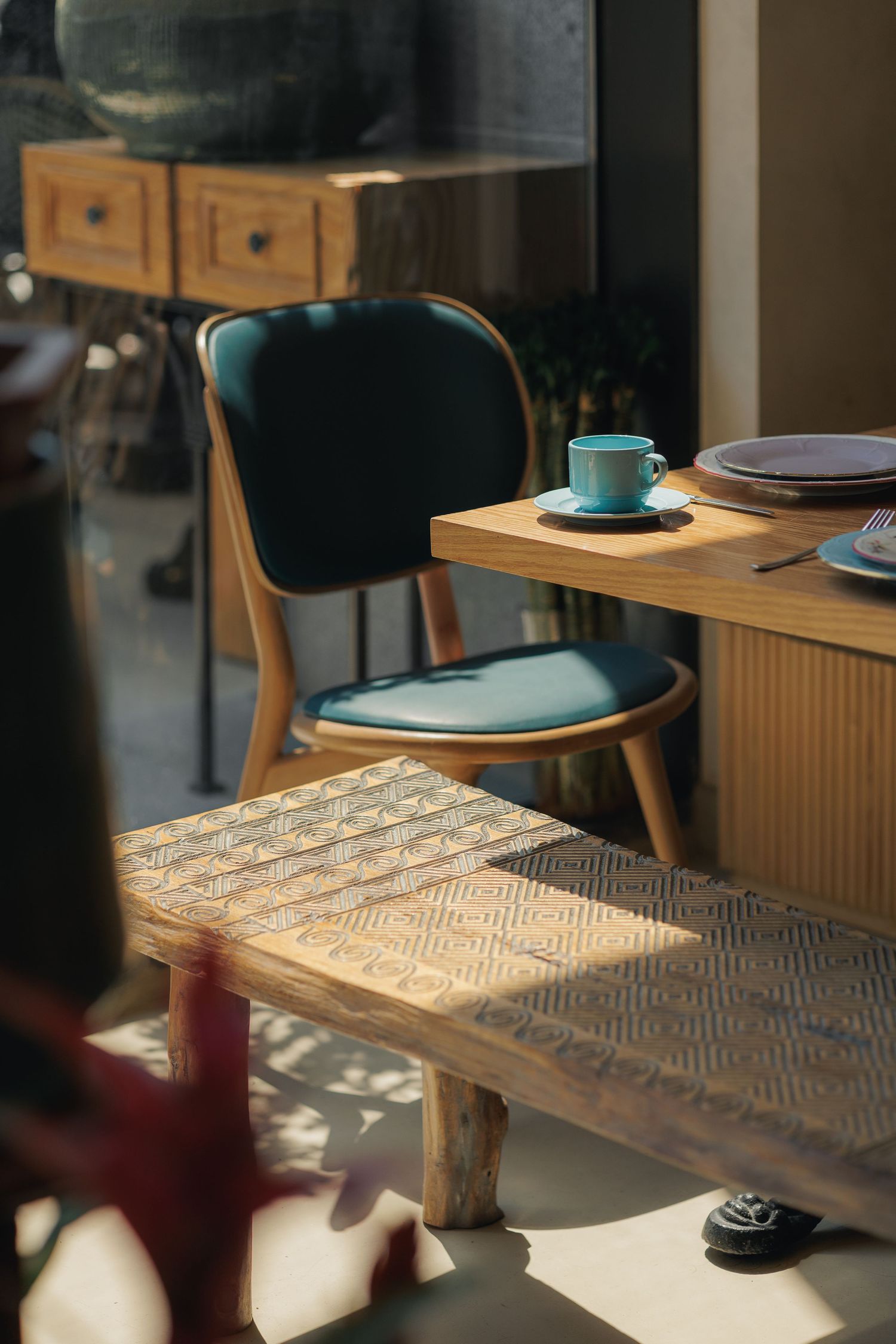 Sunlit cafe corner with teal velvet chair, wooden ribbed table, blue cup, and warm dappled light.