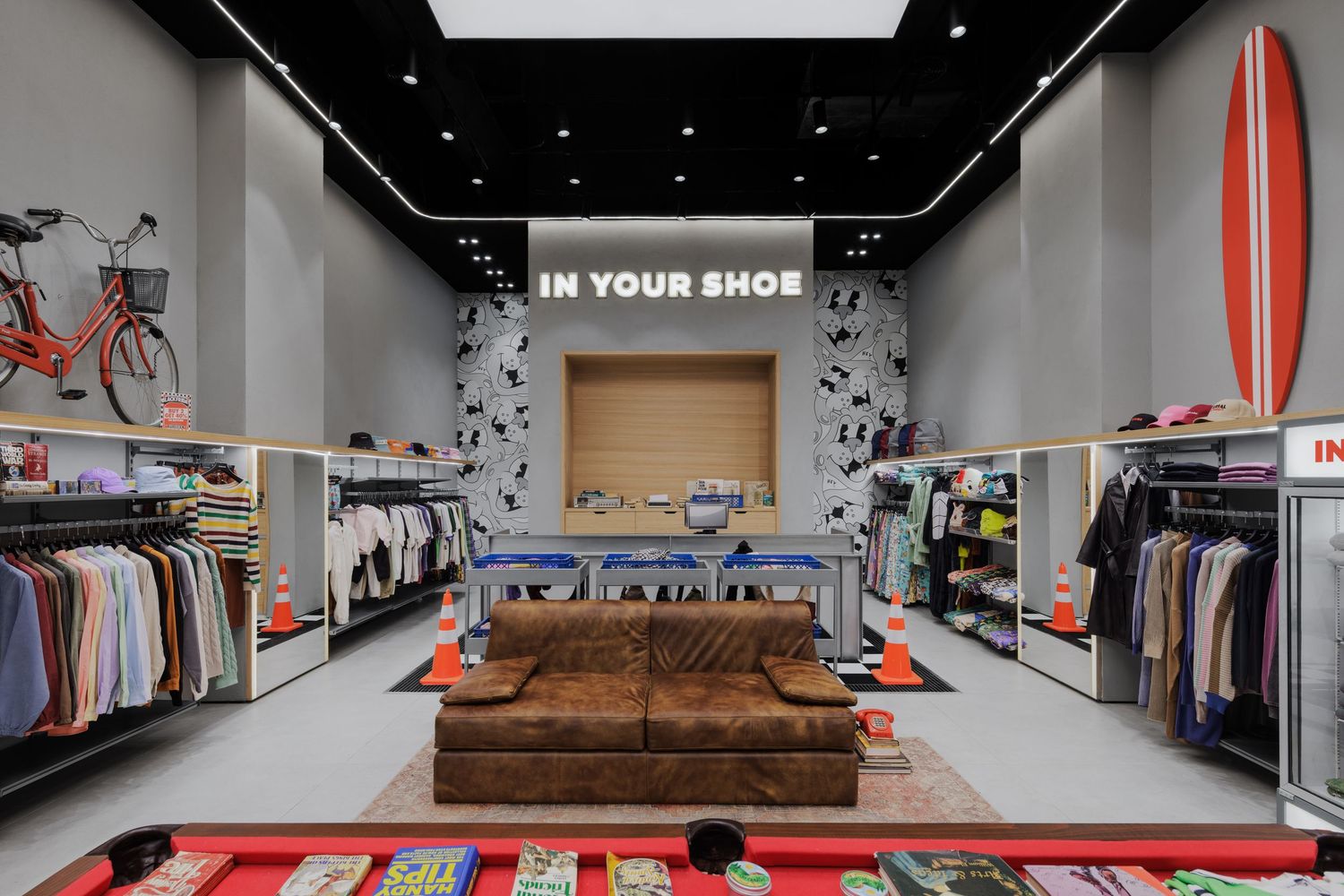 Casual retail store interior with colorful clothing racks, brown leather sofa, red striped rug, and IN YOUR SHOE signage.