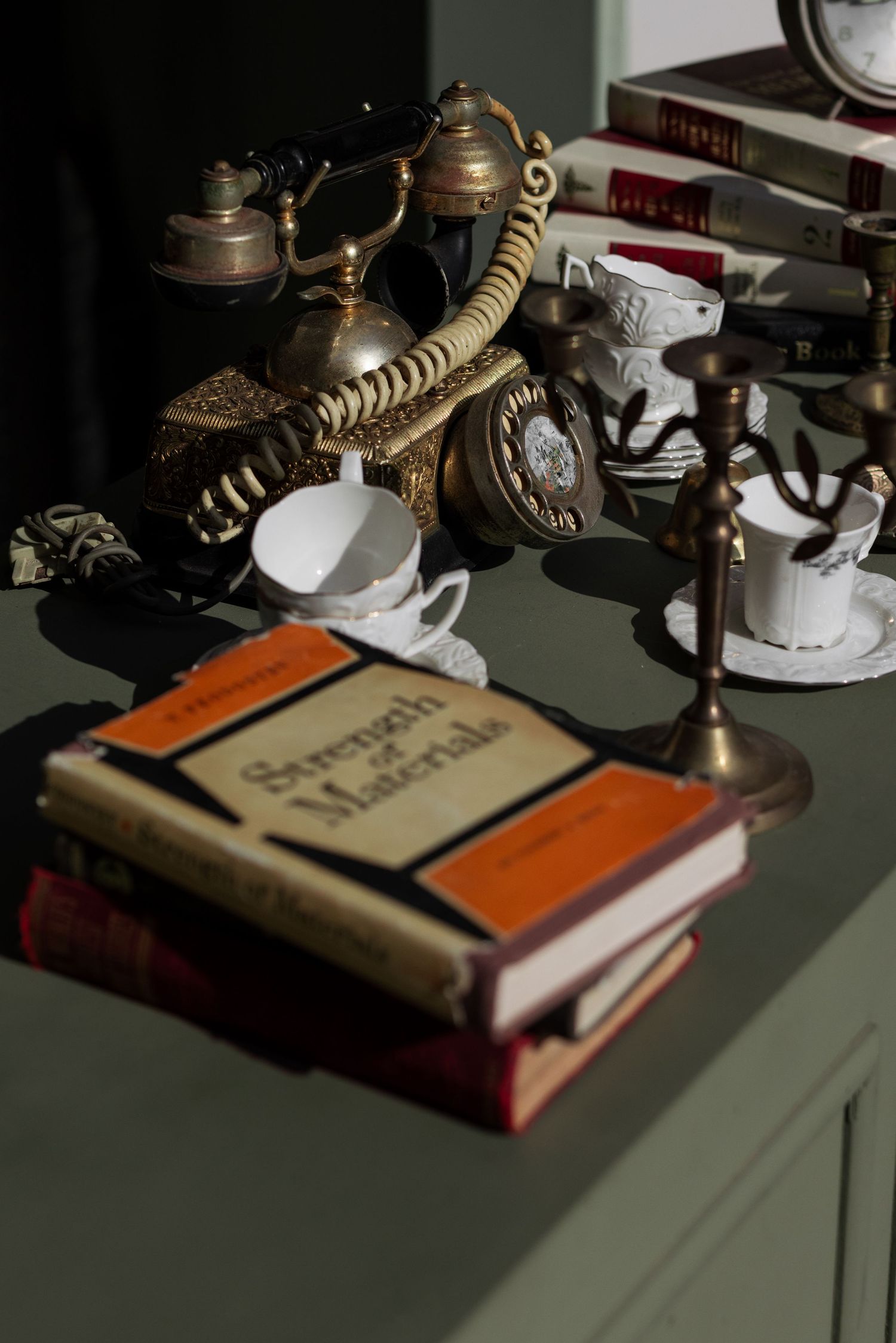 Vintage brass telephone, candlestick, and Strength of Materials books on a green dresser in warm light.