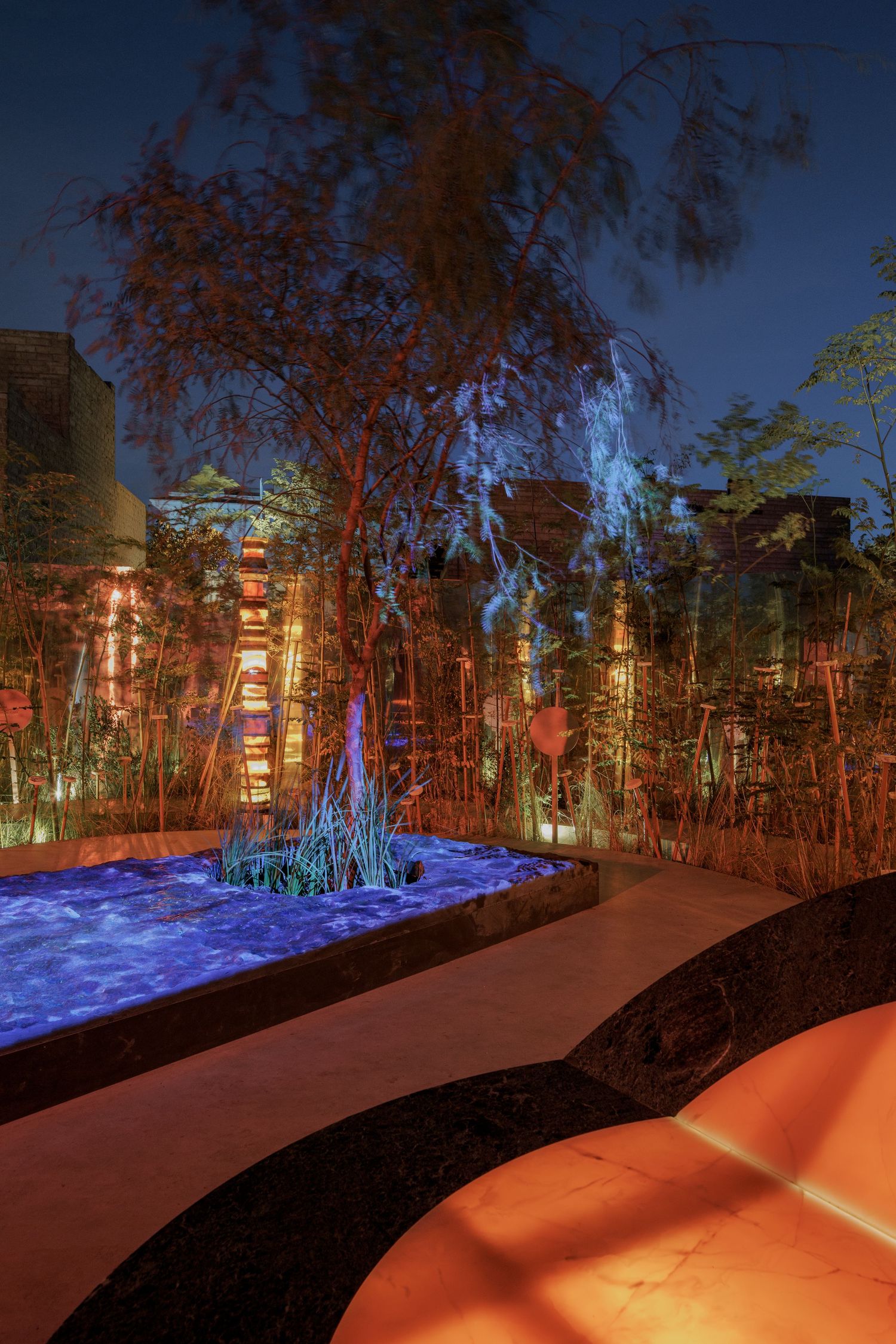 Night garden installation featuring blue glowing pool, red-lit tree, and stacked illuminated stone columns.