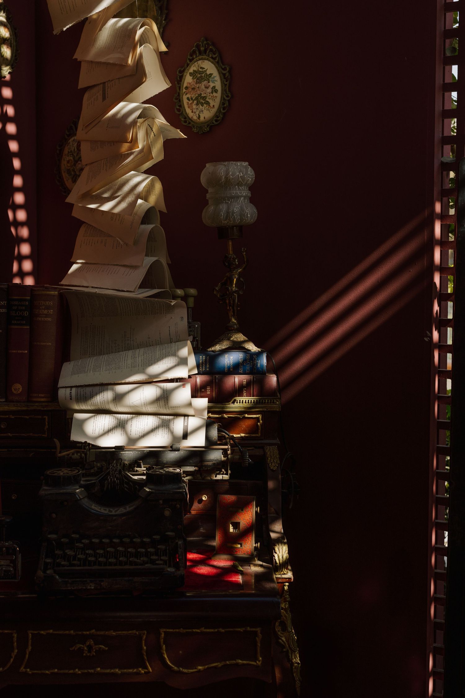 Antique typewriter with stacked open books in dramatic sunlight beside ornate lamp on dark wood desk.