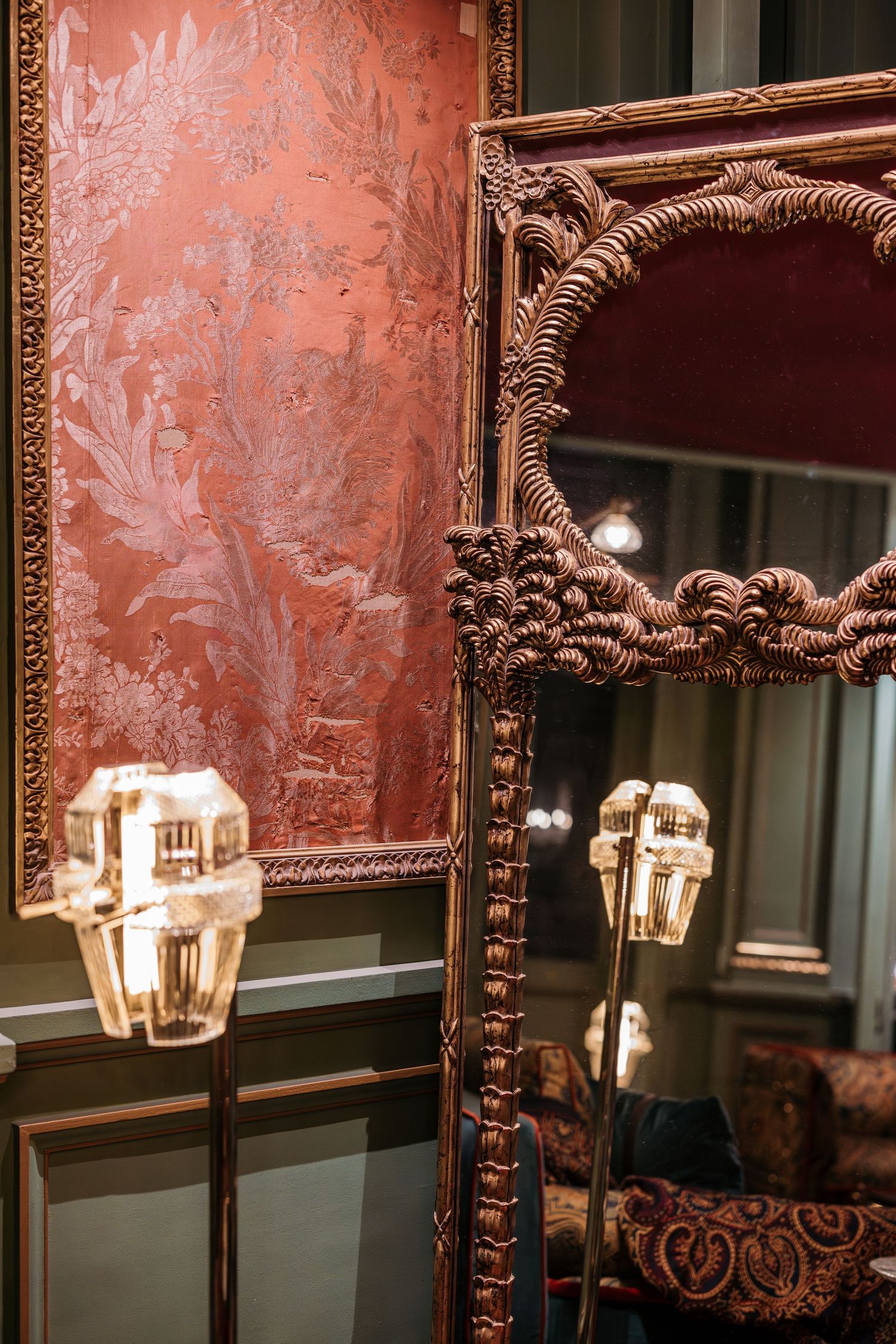Ornate gilded mirror reflecting crystal lamps against luxurious rose damask wallpaper in elegant interior.