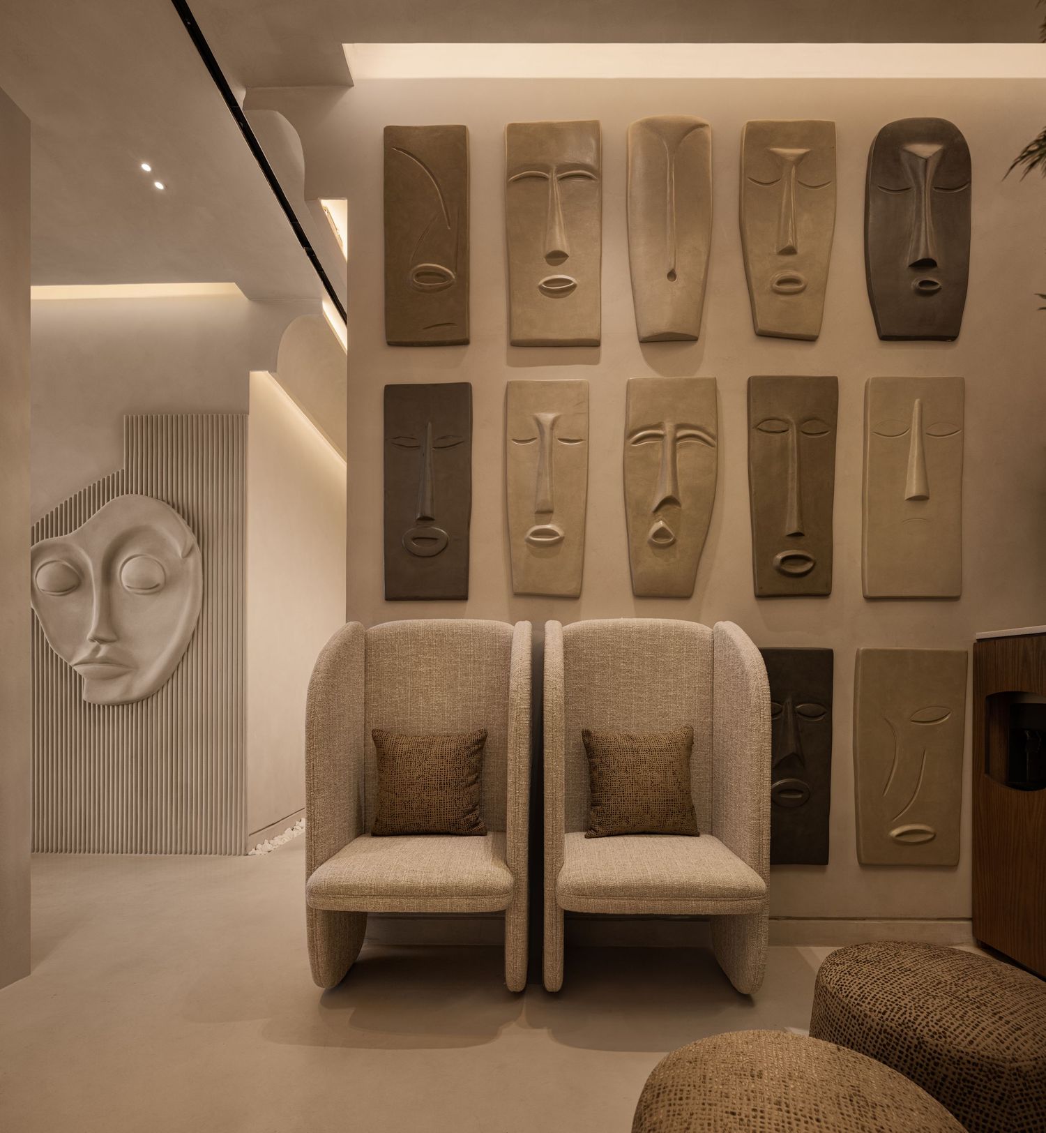 Elegant neutral-toned lounge with abstract face wall sculptures and two plush wingback chairs with cushions.