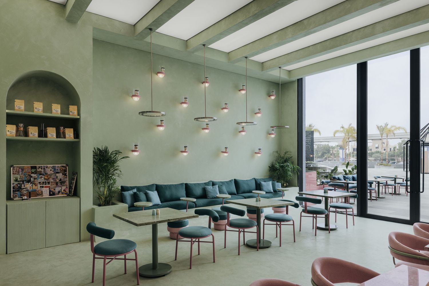 Stylish cafe with sage green walls, copper pendant lights, teal upholstered seating, and city views through large windows.