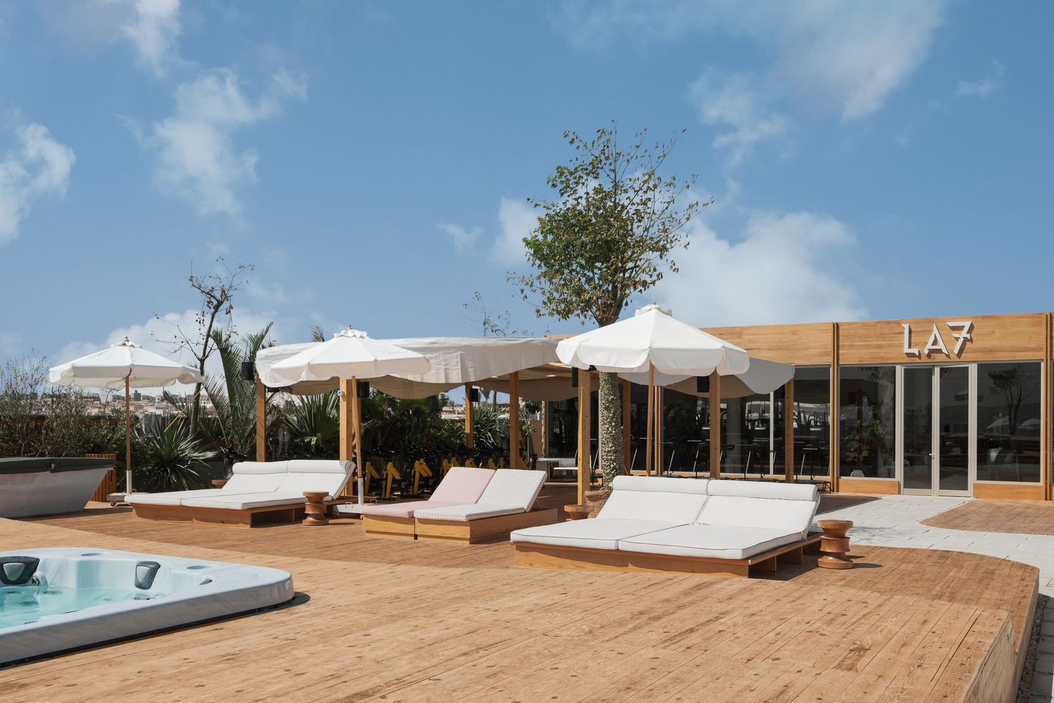 LA7 rooftop terrace with wooden deck, white lounge chairs, parasols, jacuzzi, and olive tree under blue sky.