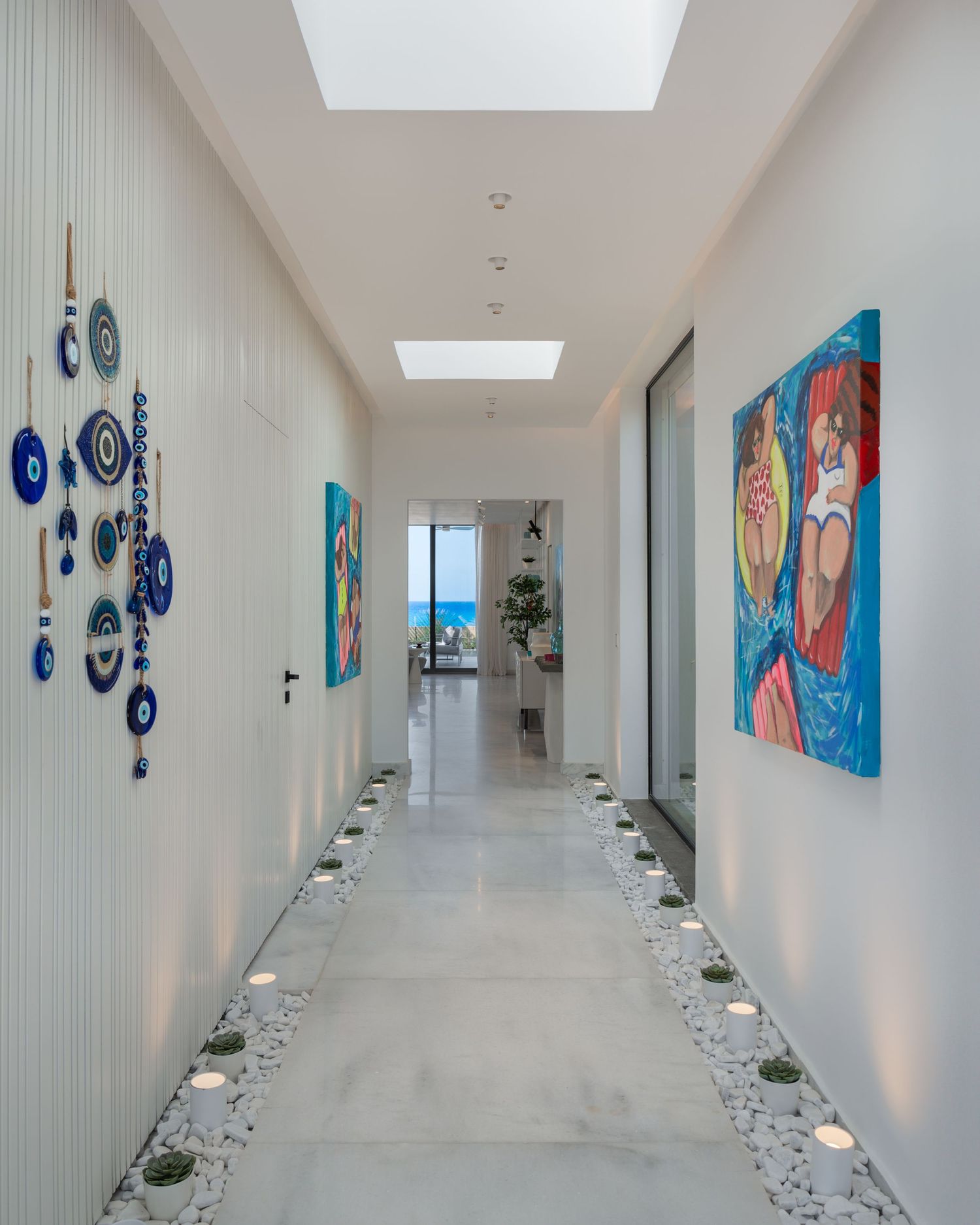 Bright modern hallway with blue evil eye wall decor, colorful artwork, candles, and ocean view at end.