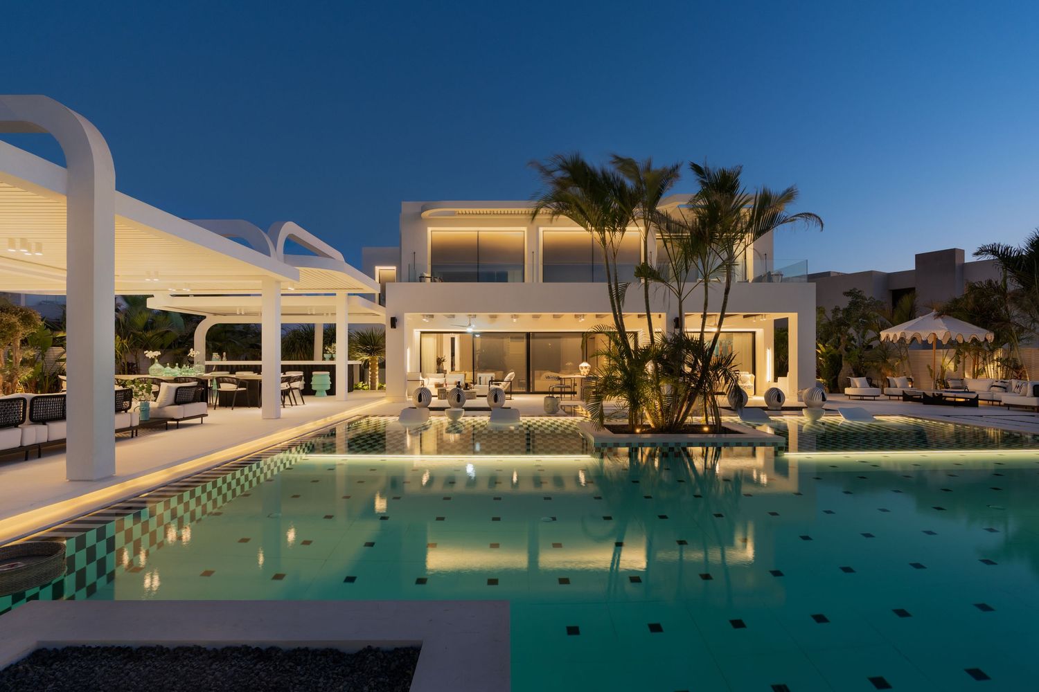 Luxurious white villa at night featuring a large illuminated pool, white pergolas, palm trees, and spacious outdoor living area.
