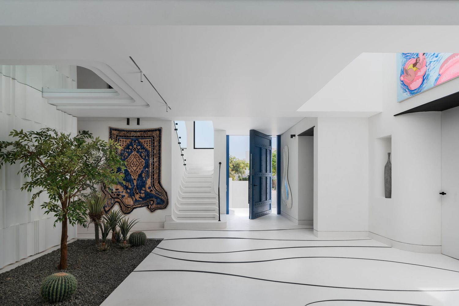 Bright white luxury villa foyer with indoor trees, ornate rug, curved staircase, and bold blue front door.