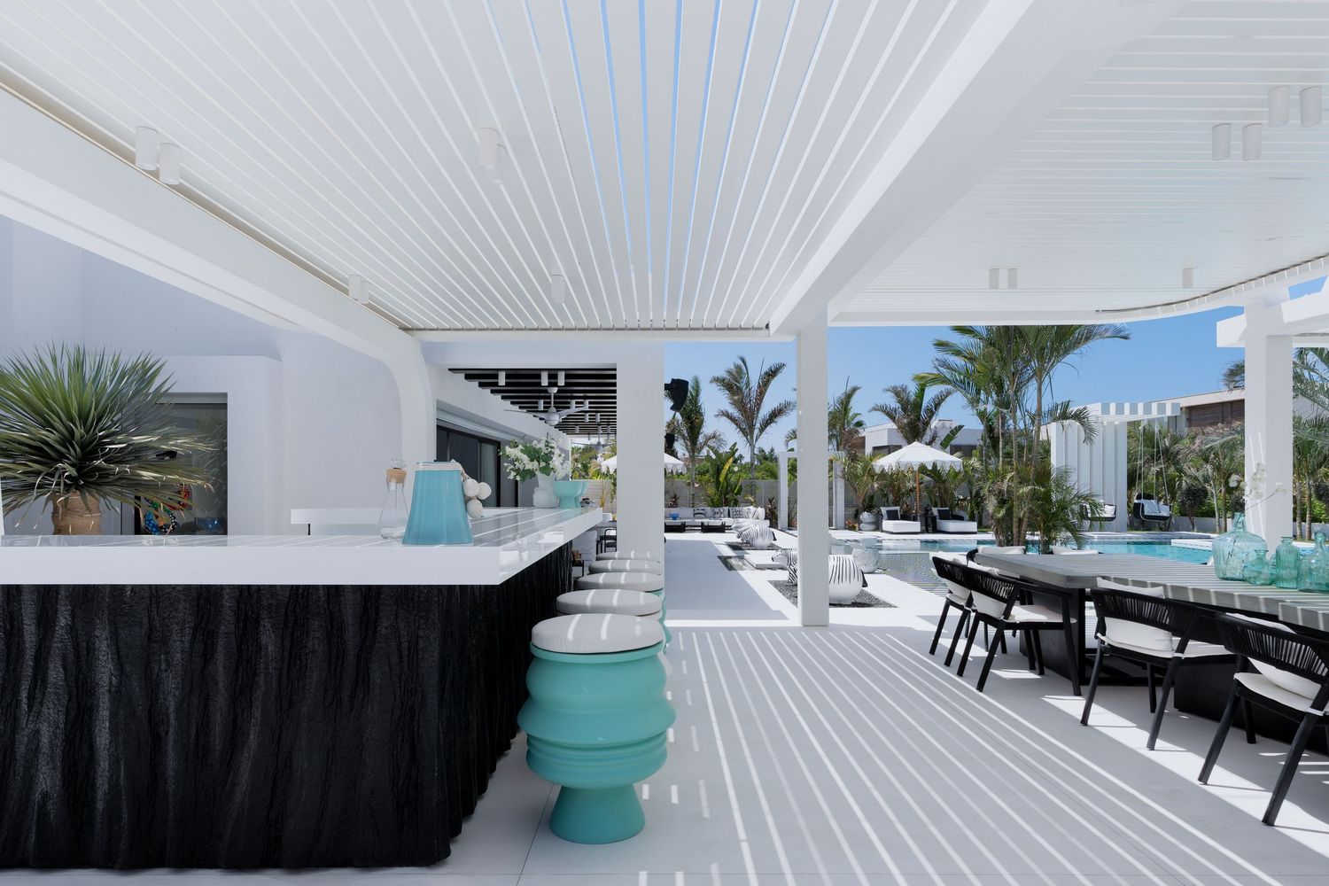 Modern white outdoor bar area with teal stools, black skirting, and poolside dining under a pergola.