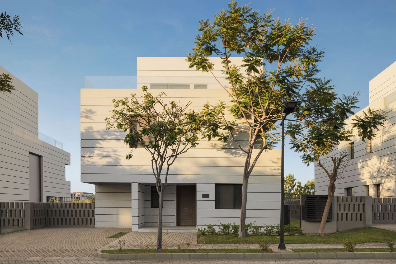 Contemporary white villa with geometric facade and young trees lining a quiet residential street.
