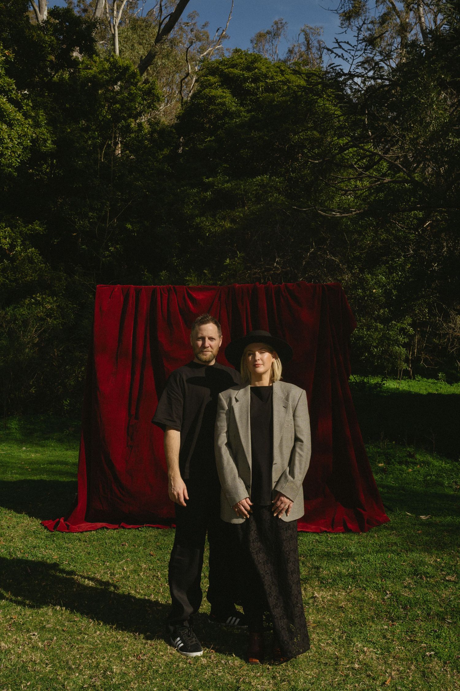 Two people stand in front of a red velvet curtain backdrop in an outdoor setting with lush greenery.