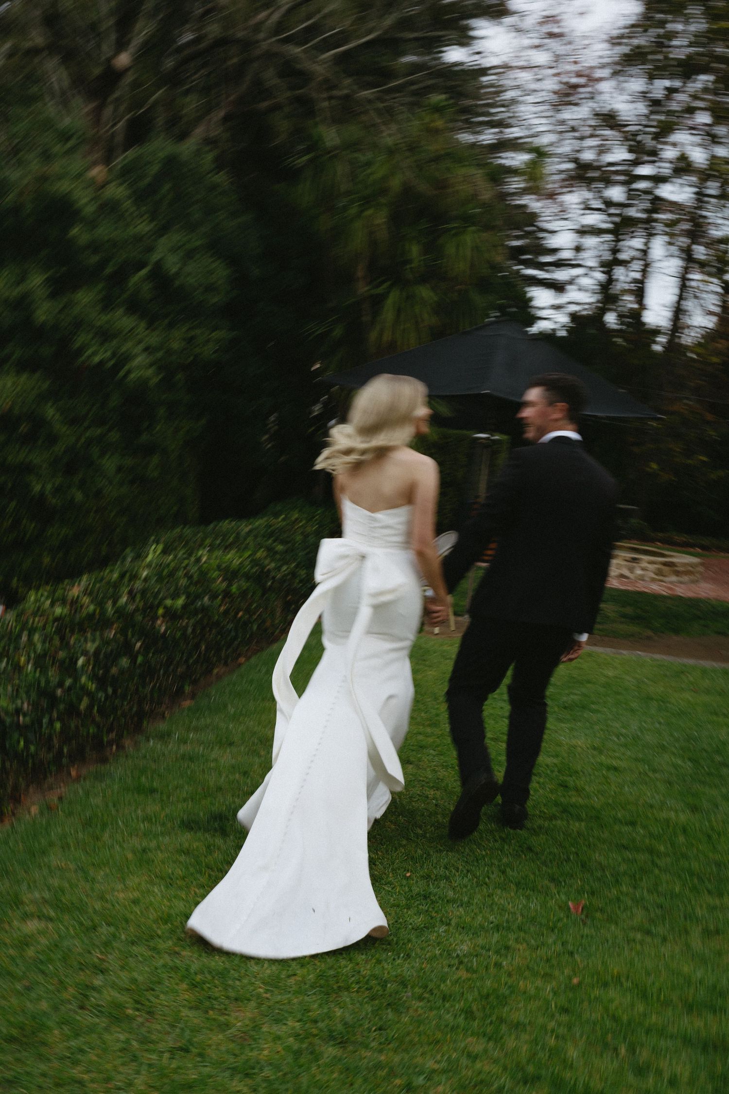 A couple in wedding attire dance together on a green lawn surrounded by trees and gardens.