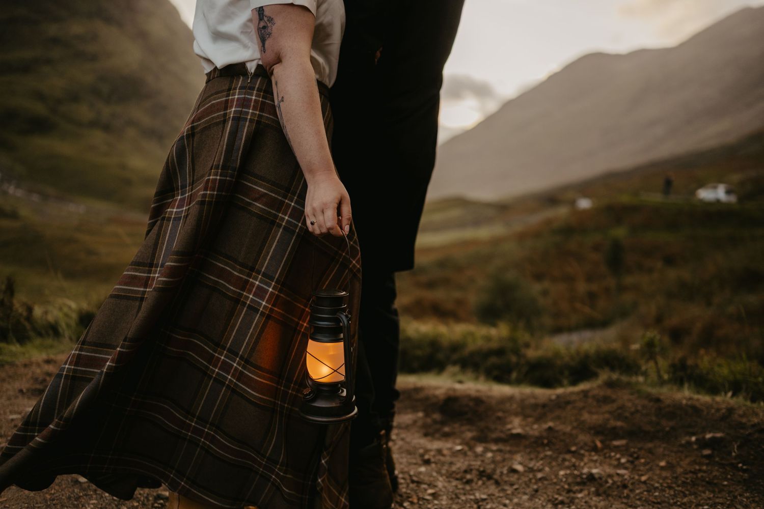 A glowing lantern held against a plaid skirt in a moody Scottish Highland landscape at dusk.