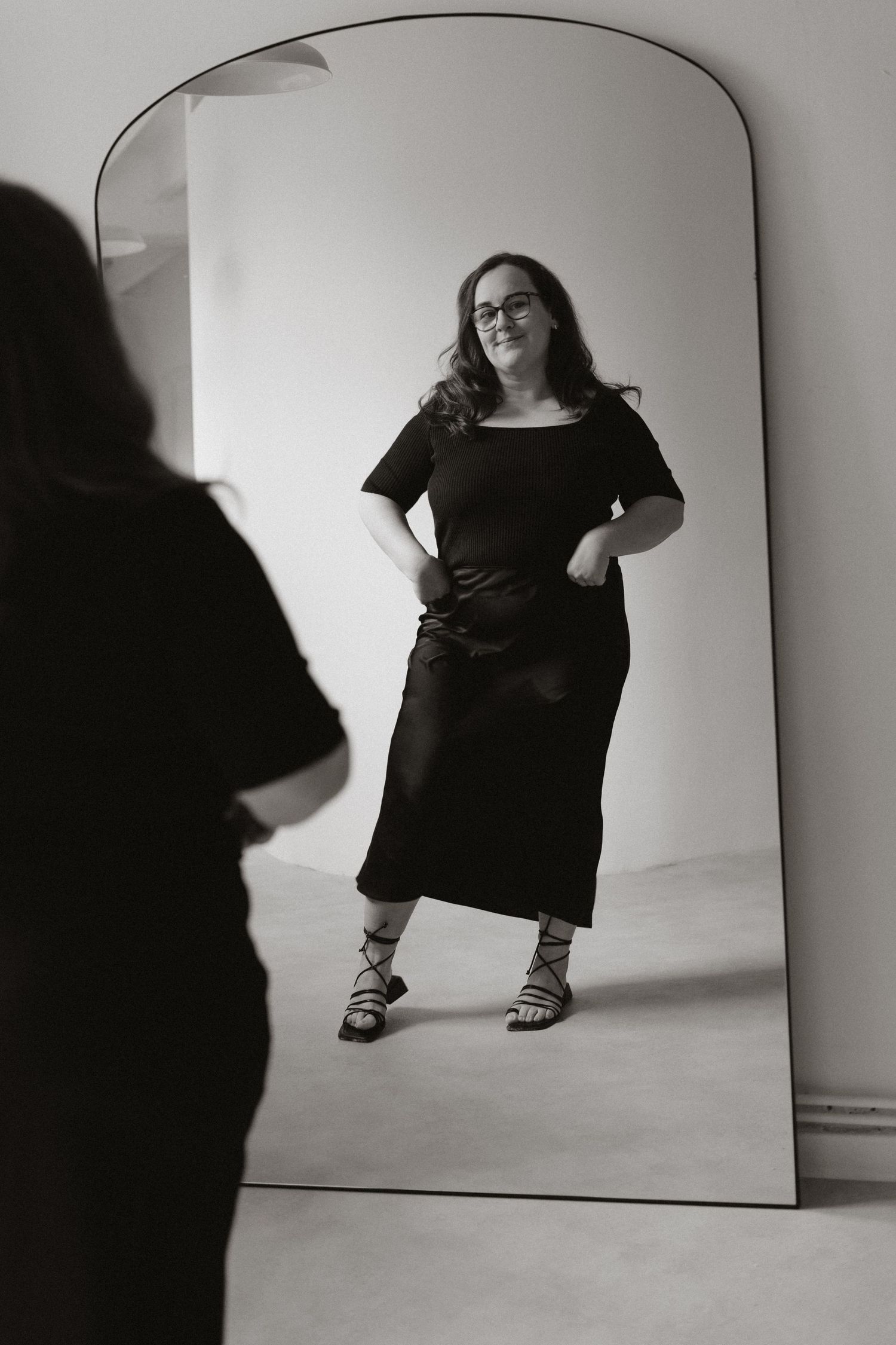 Artistic black and white fashion photography showing someone in a dark dress against a minimalist white studio backdrop.