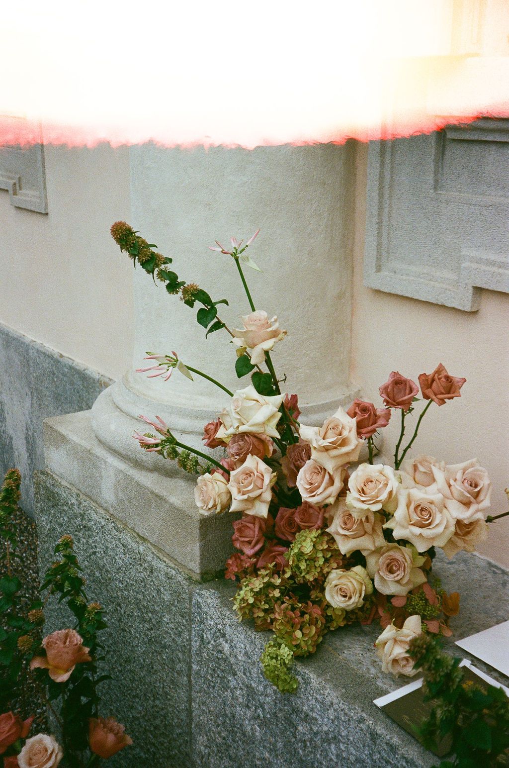 Elegant floral arrangement of blush and dusty rose roses with greenery displayed on a stone pedestal outdoors.