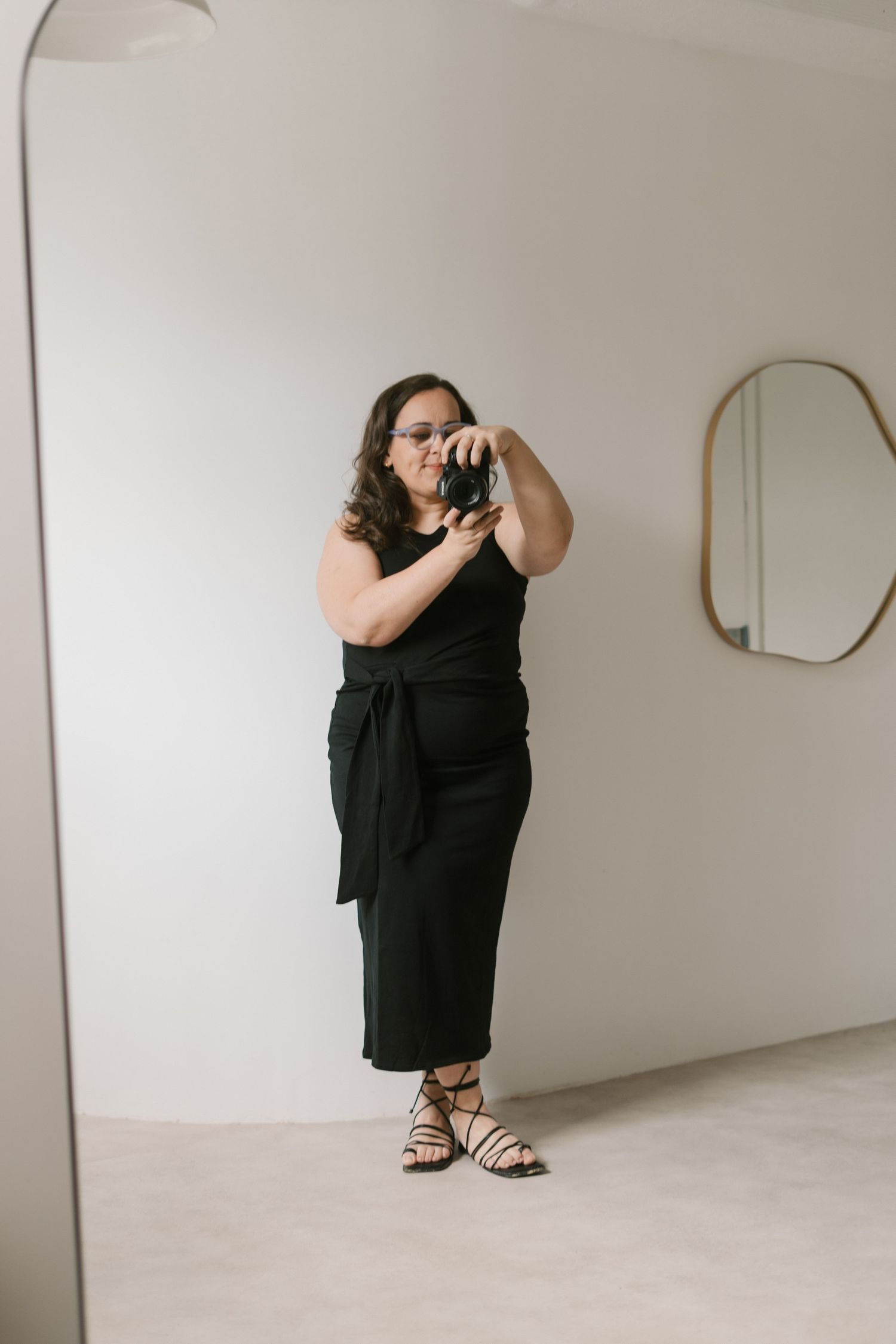 Someone wearing a black jumpsuit takes a mirror selfie in a minimalist room with white walls and an oval mirror.