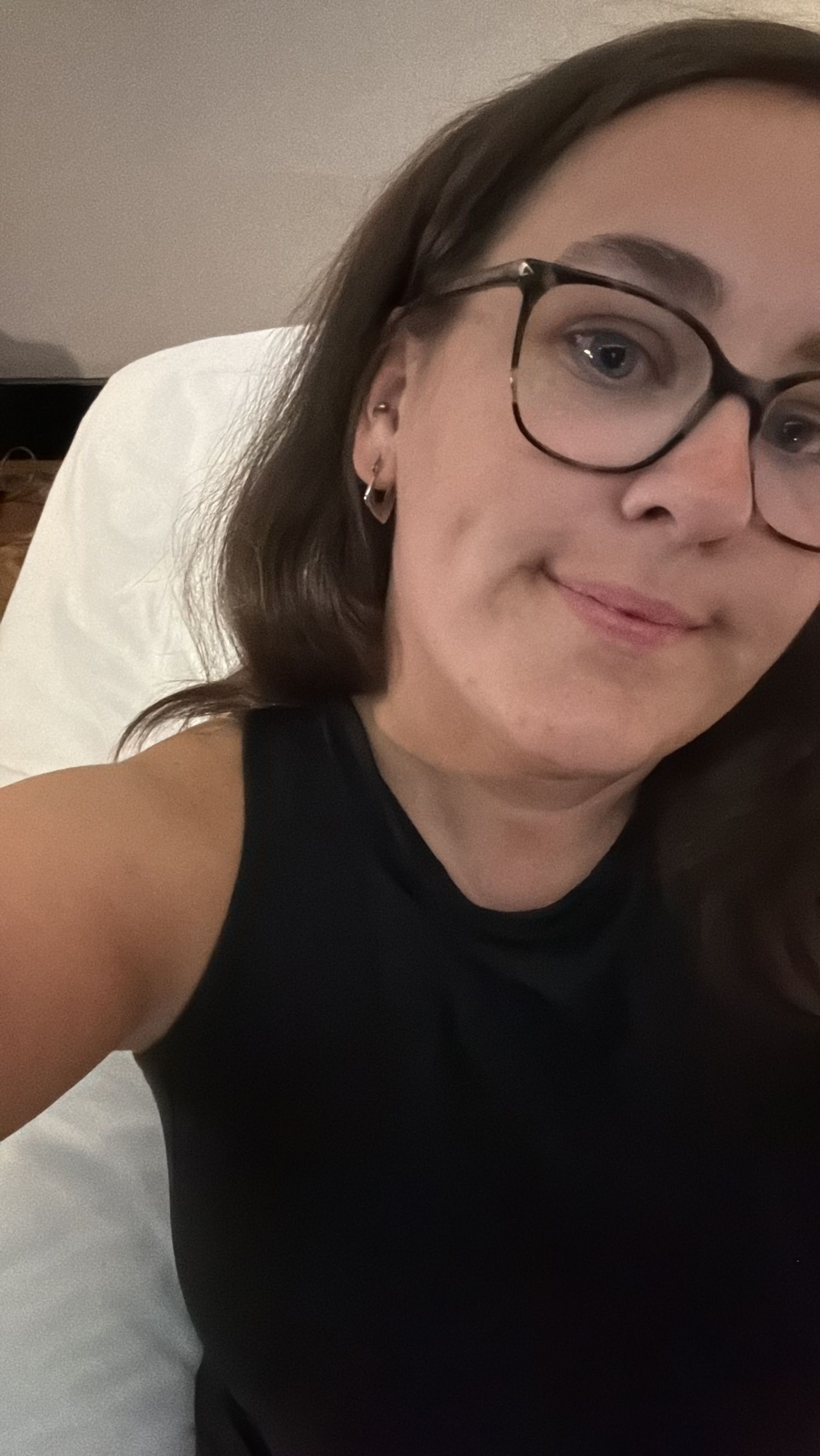 Young woman with dark hair and black glasses taking a selfie, wearing a black sleeveless top, smiling softly.