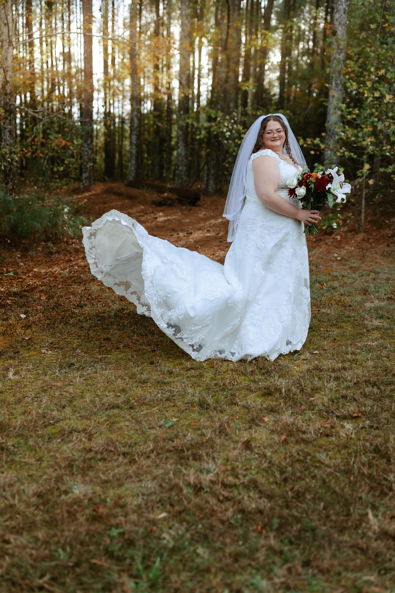 Bride in white lace gown with flowing train and veil holds bouquet in autumn forest setting.