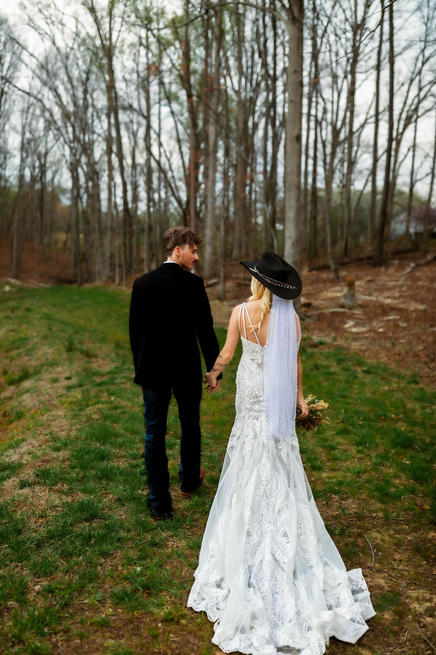 A couple holding hands in wedding attire walks through a wooded area with bare trees and green grass.