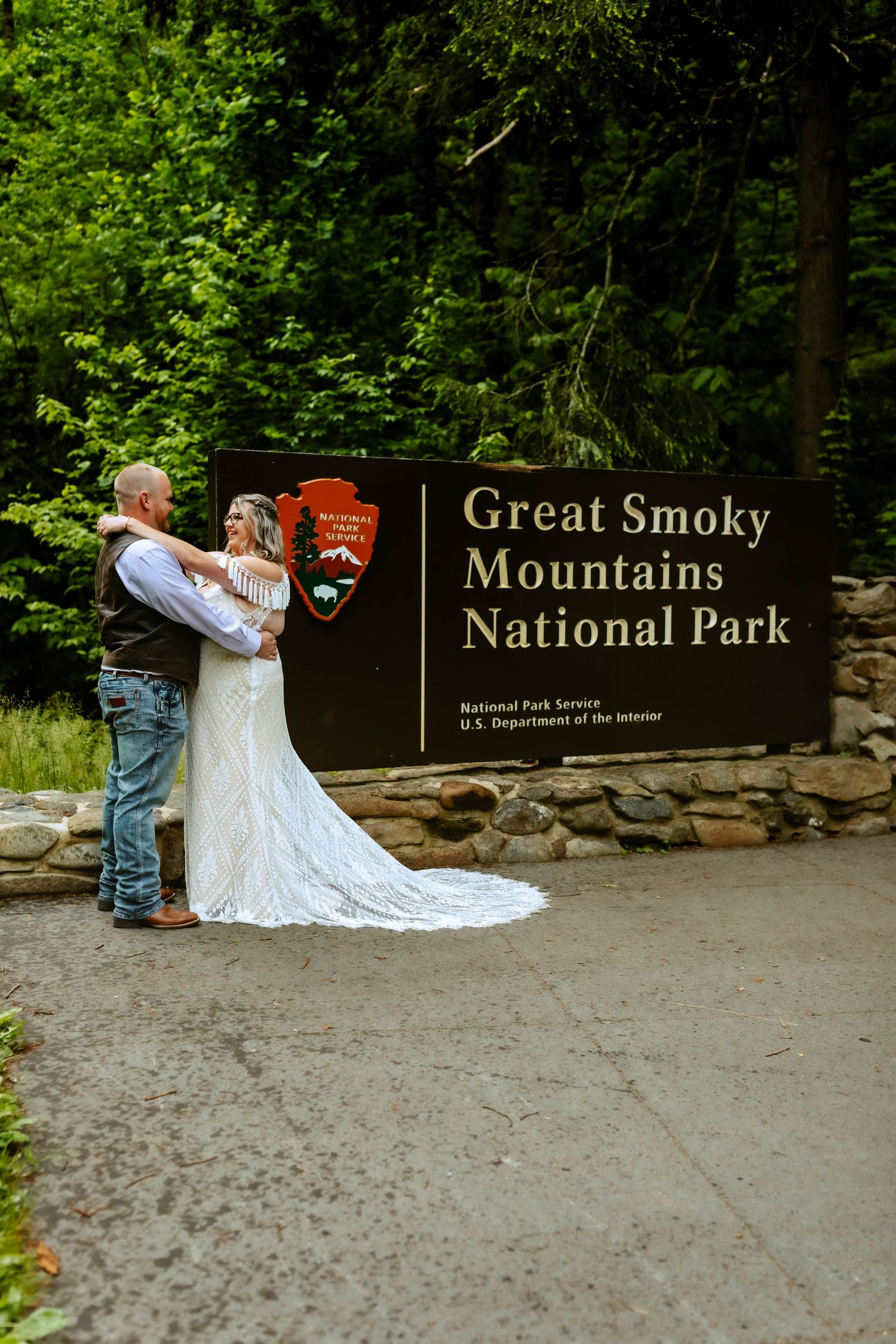 A couple embraces in front of the Great Smoky Mountains National Park entrance sign on a lush forest path.