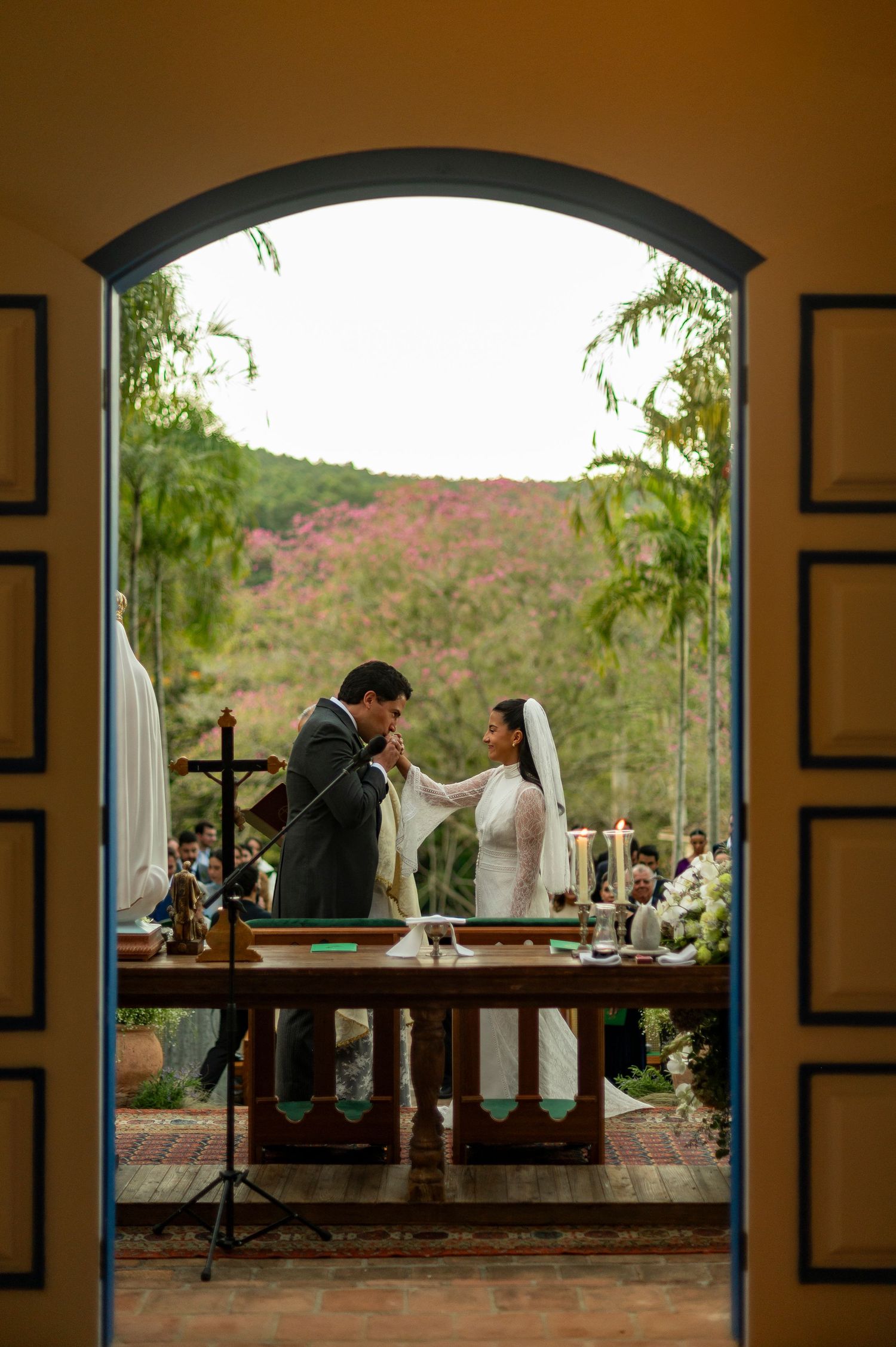 Bride and groom exchange vows at outdoor wedding ceremony framed by arched doorway with lush tropical greenery backdrop.