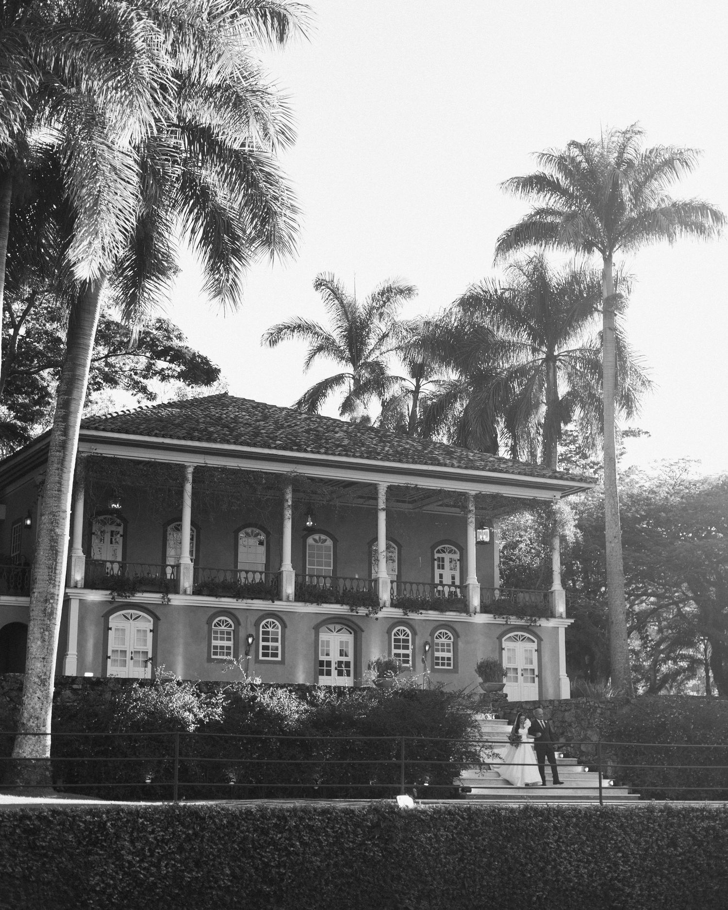 Black and white photo of a colonial-style mansion with wrap-around balcony surrounded by tall palm trees.
