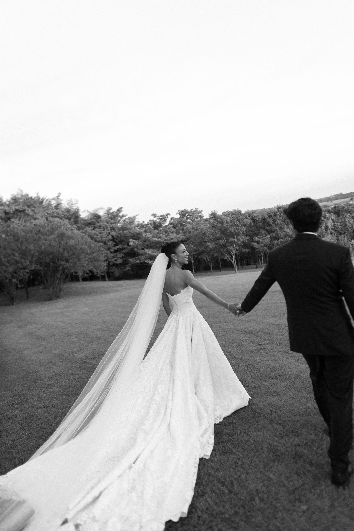 A couple in wedding attire walks together on a grassy lawn with trees in background as her long veil trails behind them.