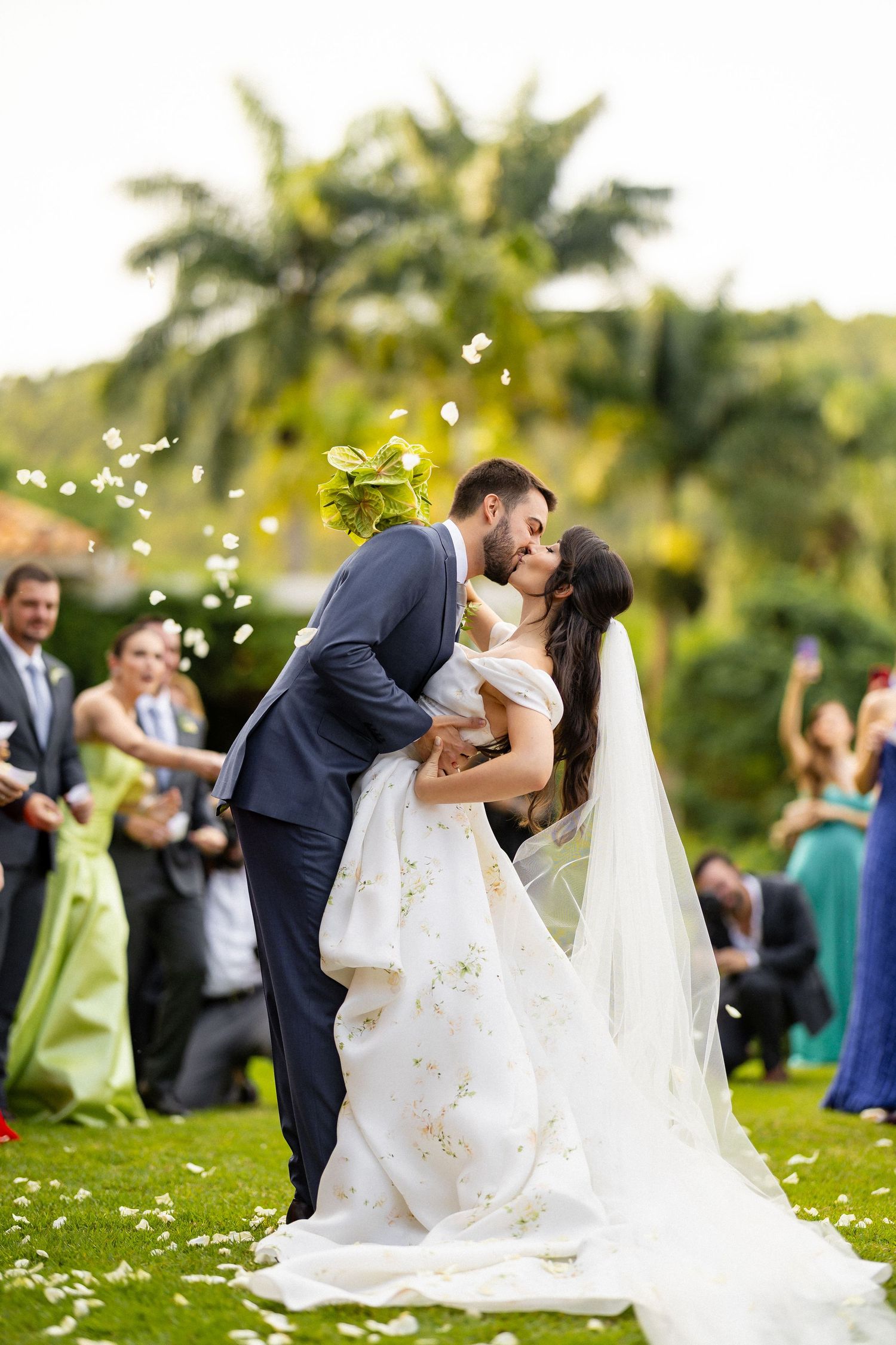 A romantic wedding scene under palm trees with flower petals floating through the air during a joyful outdoor ceremony.