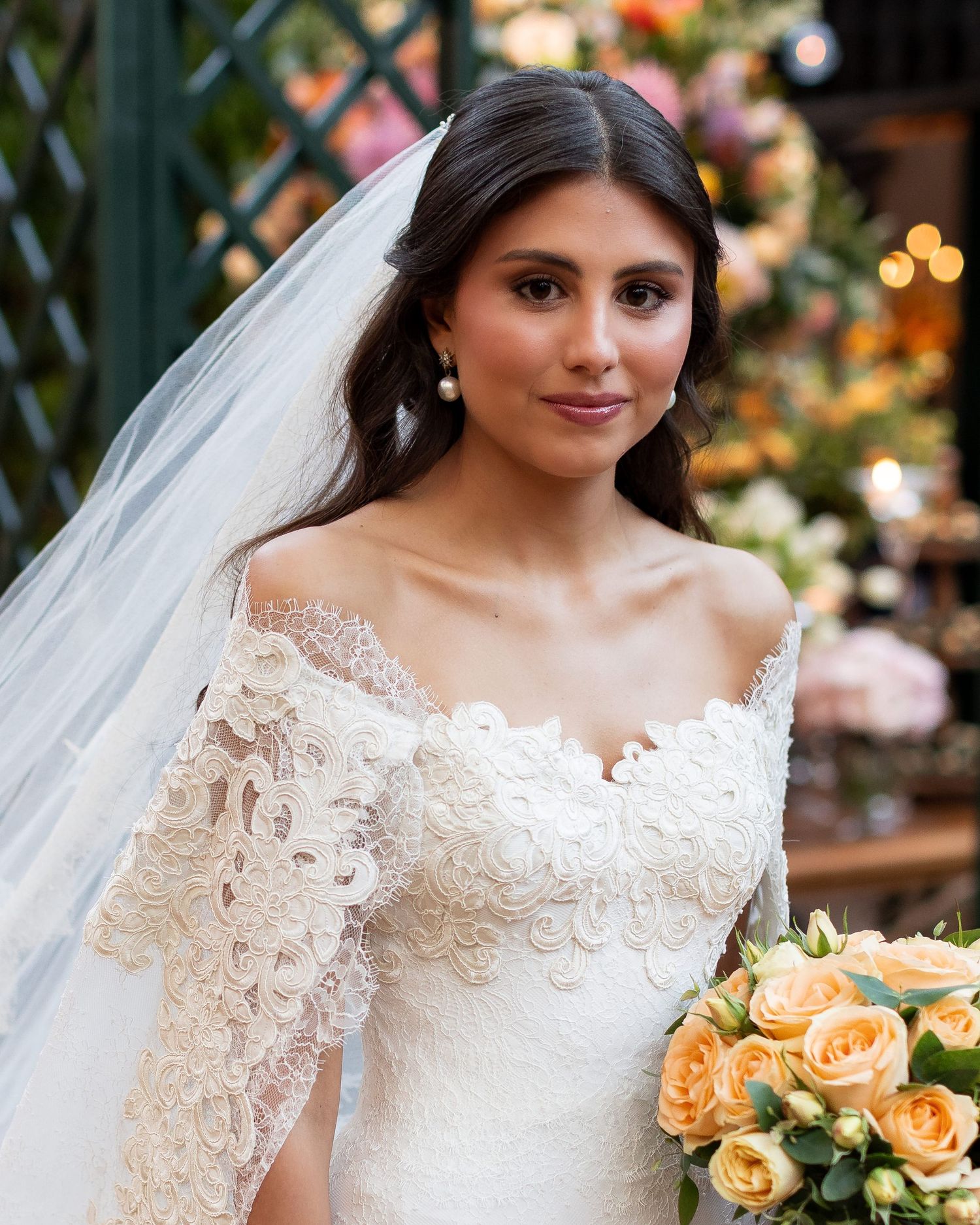 Bride wearing an elegant lace wedding gown with long sleeves and veil poses with peach-colored bouquet.