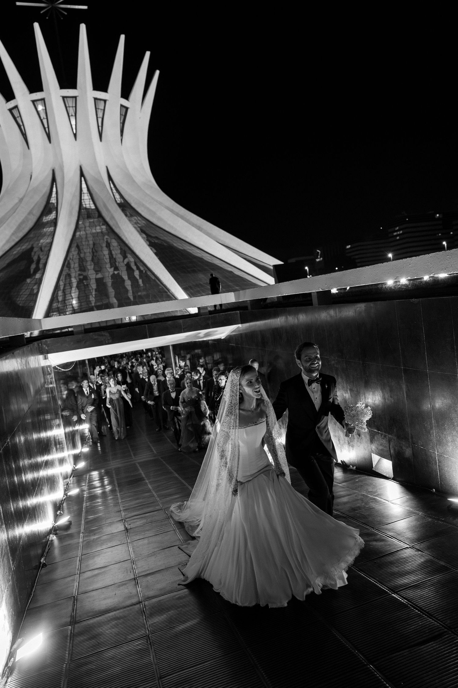 Black and white wedding photo of bride and groom walking at night outside a modern cathedral.