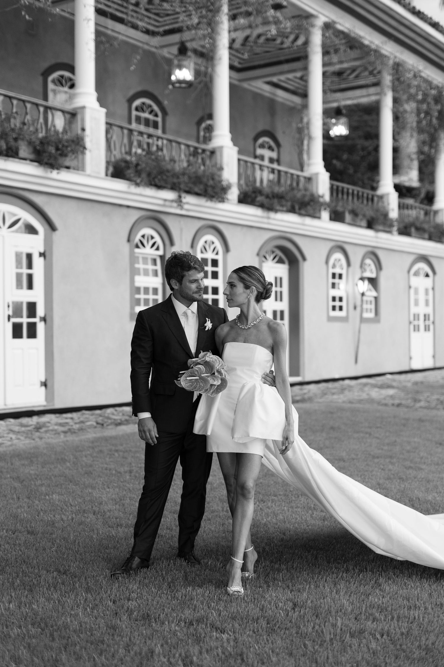 A couple poses in formal attire on a lawn in front of a historic building with classical architecture.