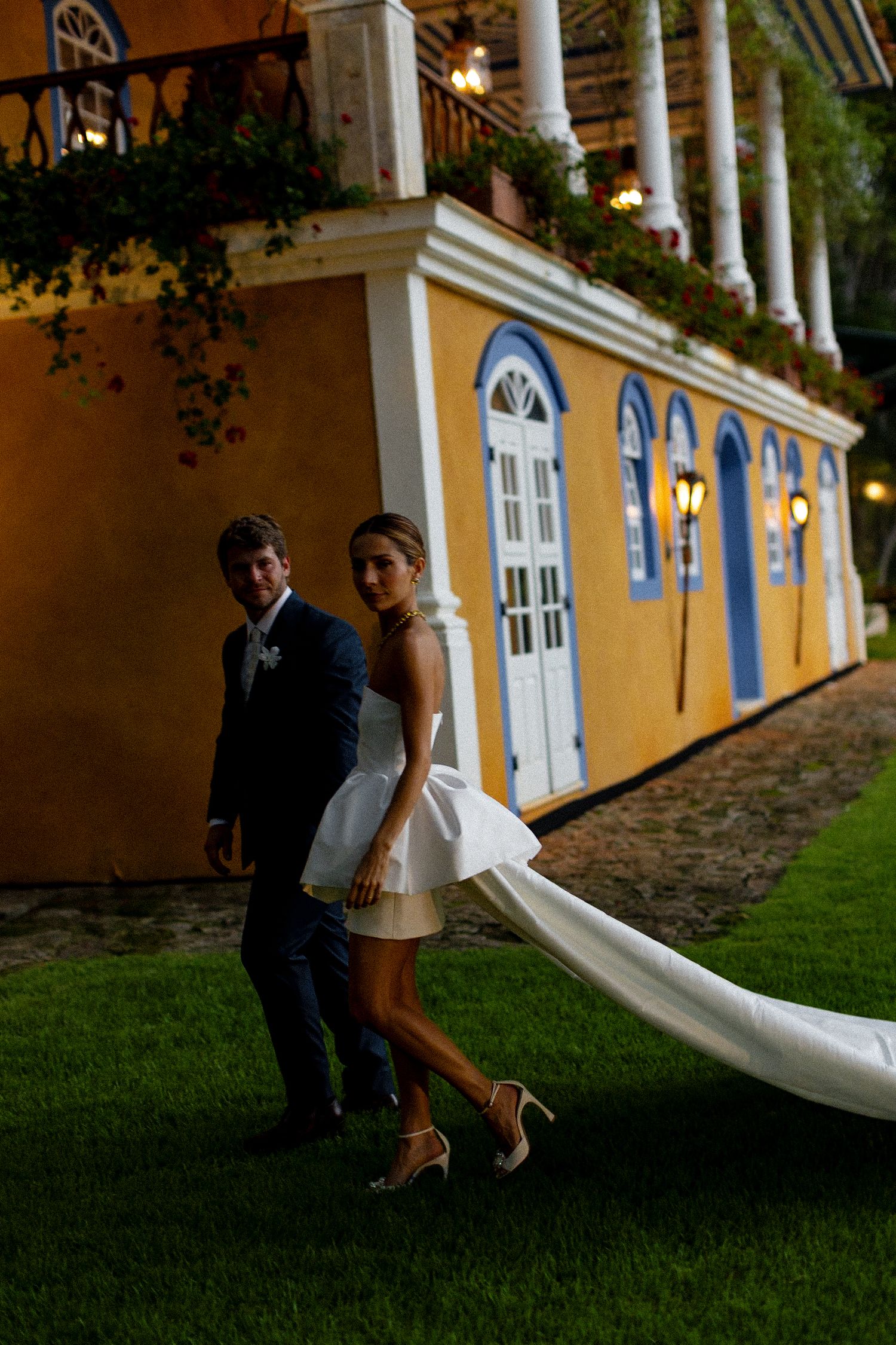 A couple in wedding attire walks on grass in front of a yellow colonial style building with white columns at dusk.