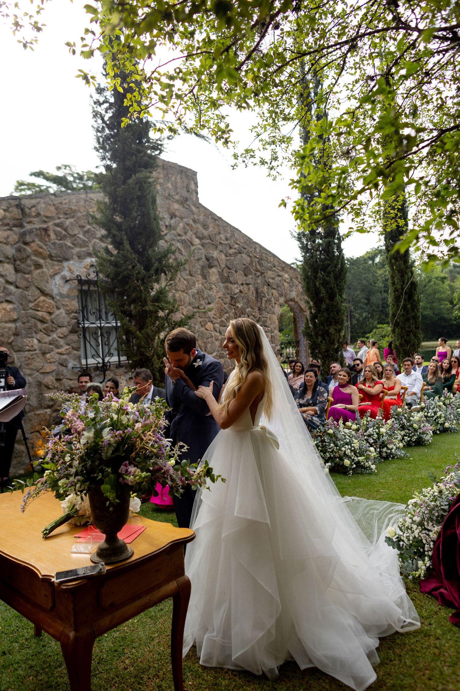 A romantic outdoor wedding ceremony takes place in front of a rustic stone chapel surrounded by lush greenery and flowers.