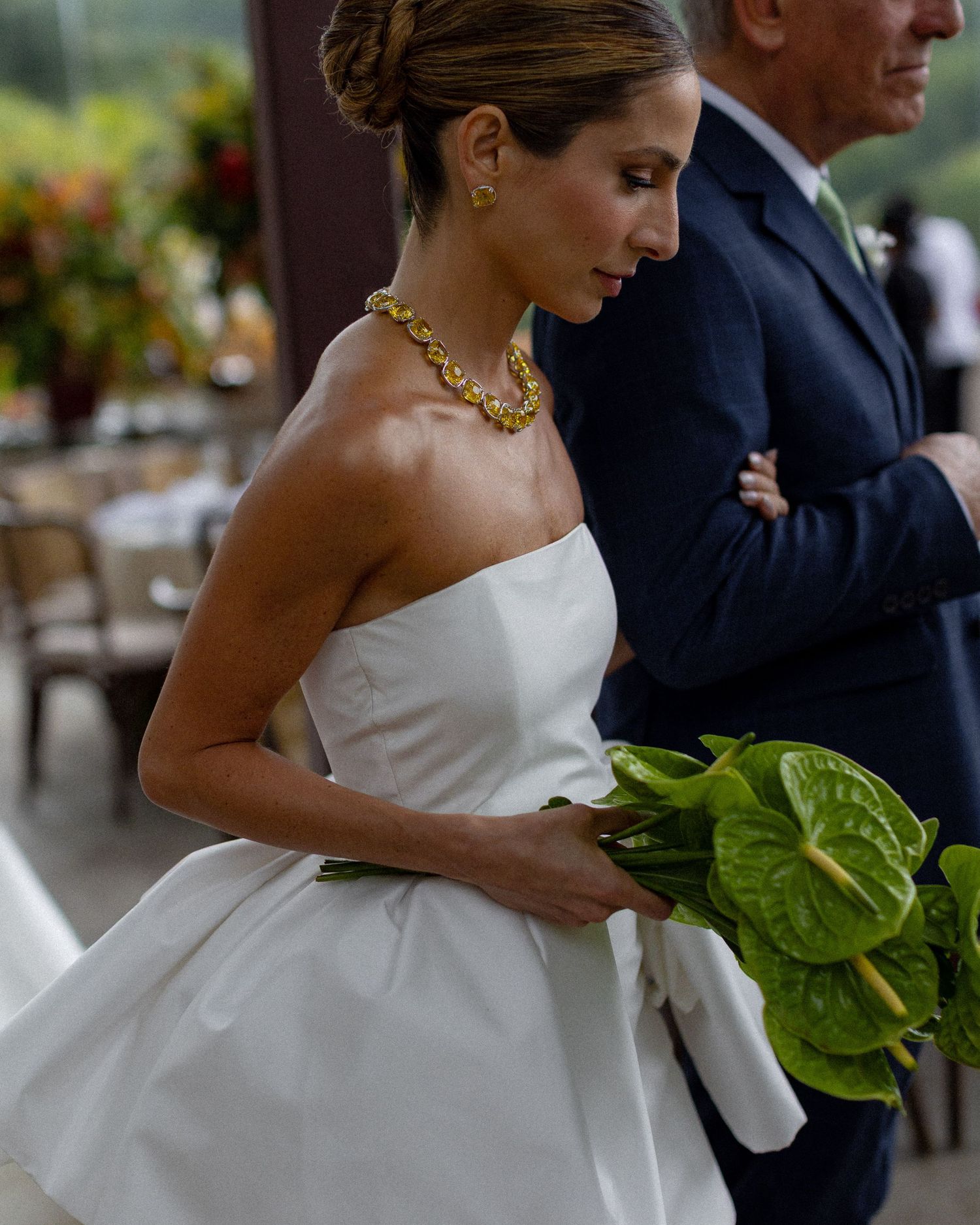 Couple in formal wedding attire pose together while holding green tropical leaves during an outdoor reception.